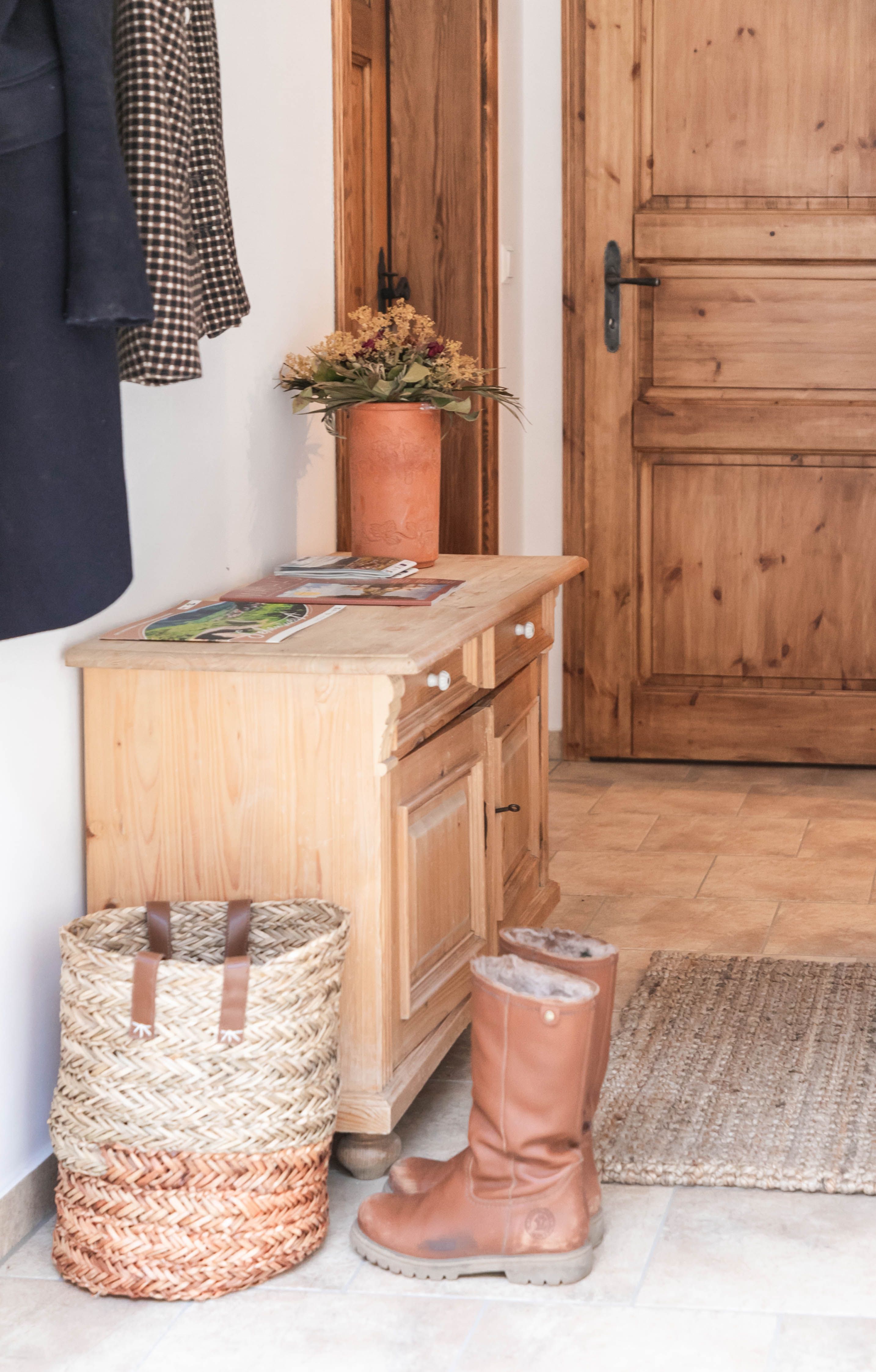 An anteroom with wooden furniture, a basket, boots and a vase of flowers.