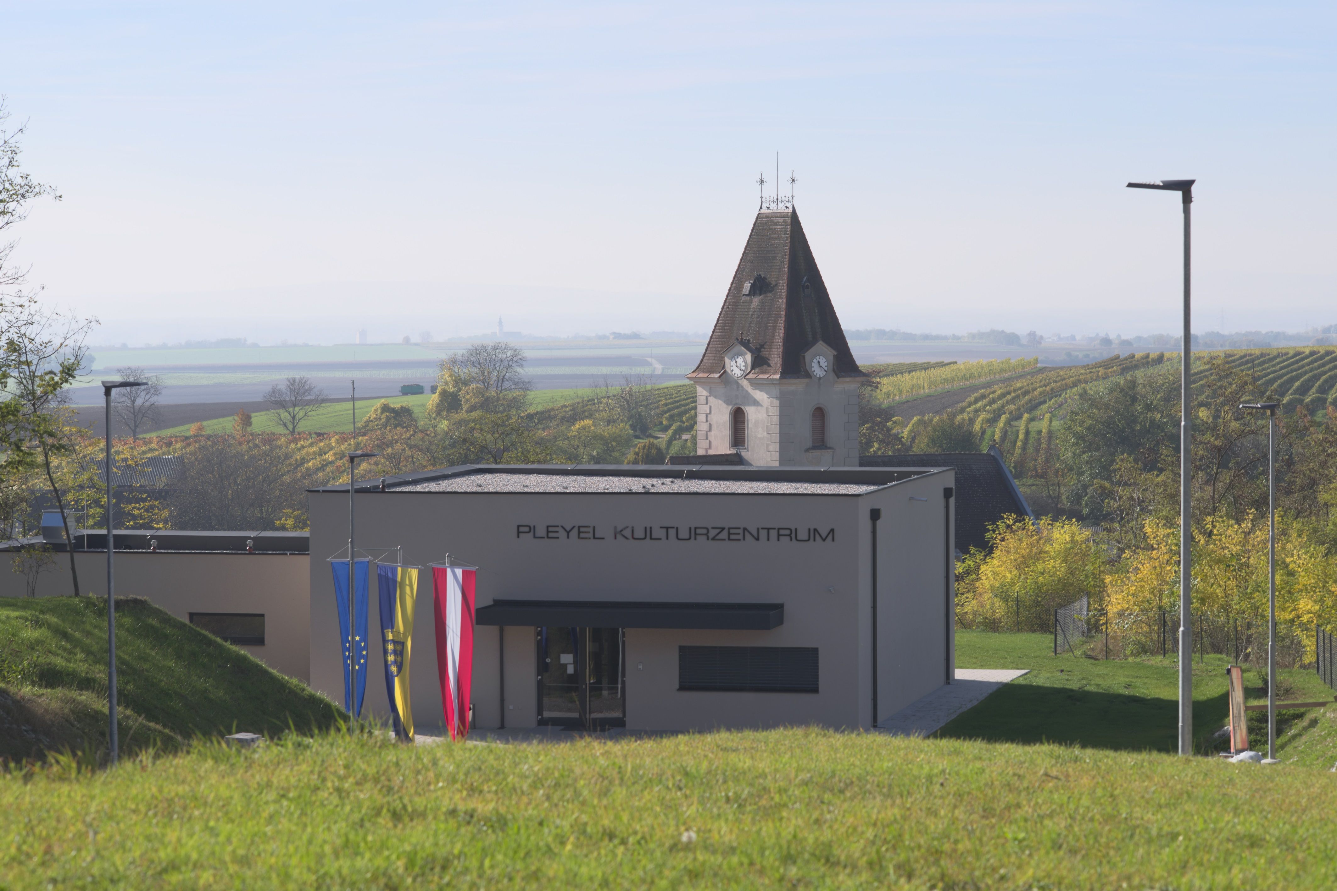 A modern building with the inscription 'Pleyel Cultural Center' in front of a rural landscape with vineyards and a church tower.