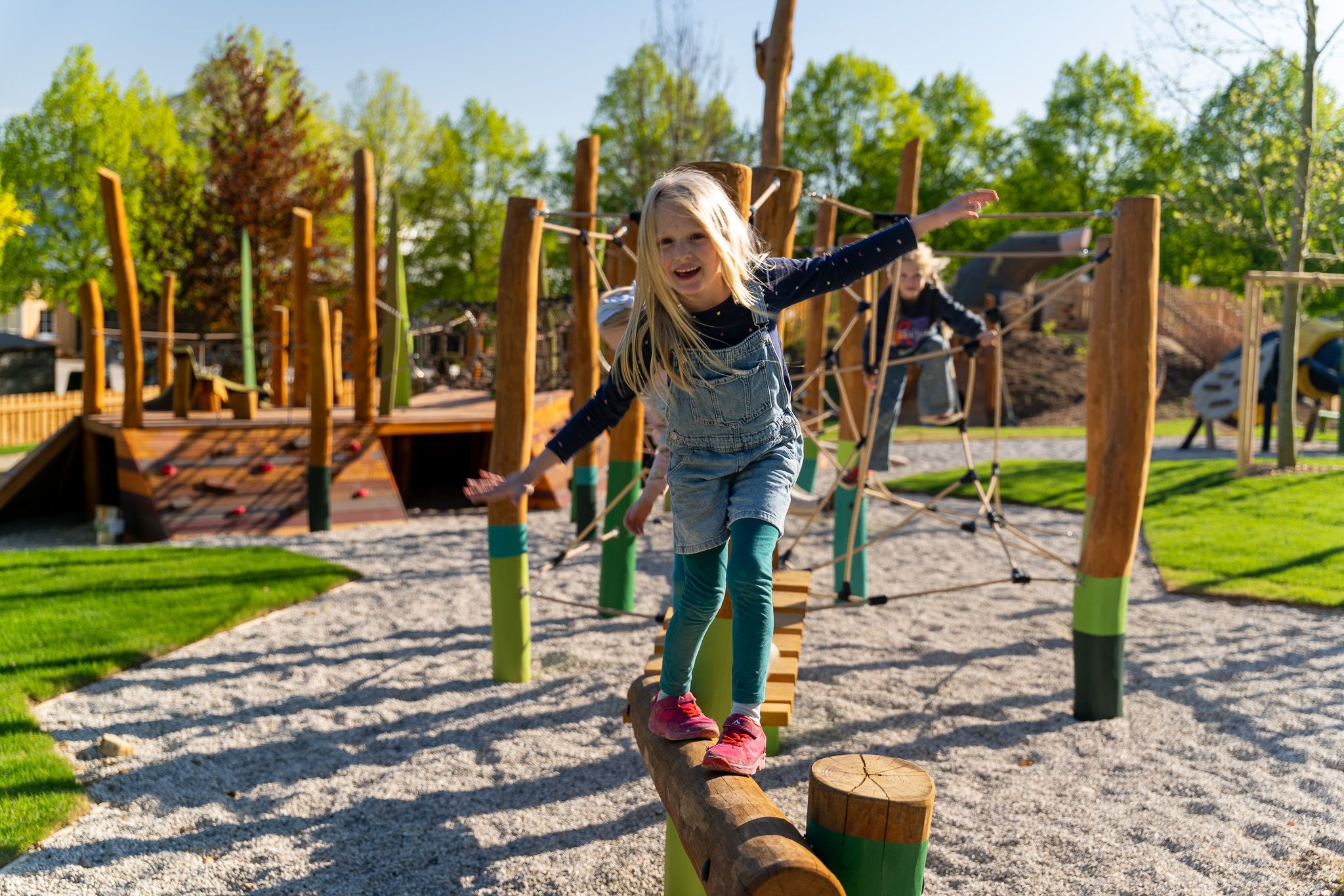 A child balances on a log in a playground with climbing frames in the background.