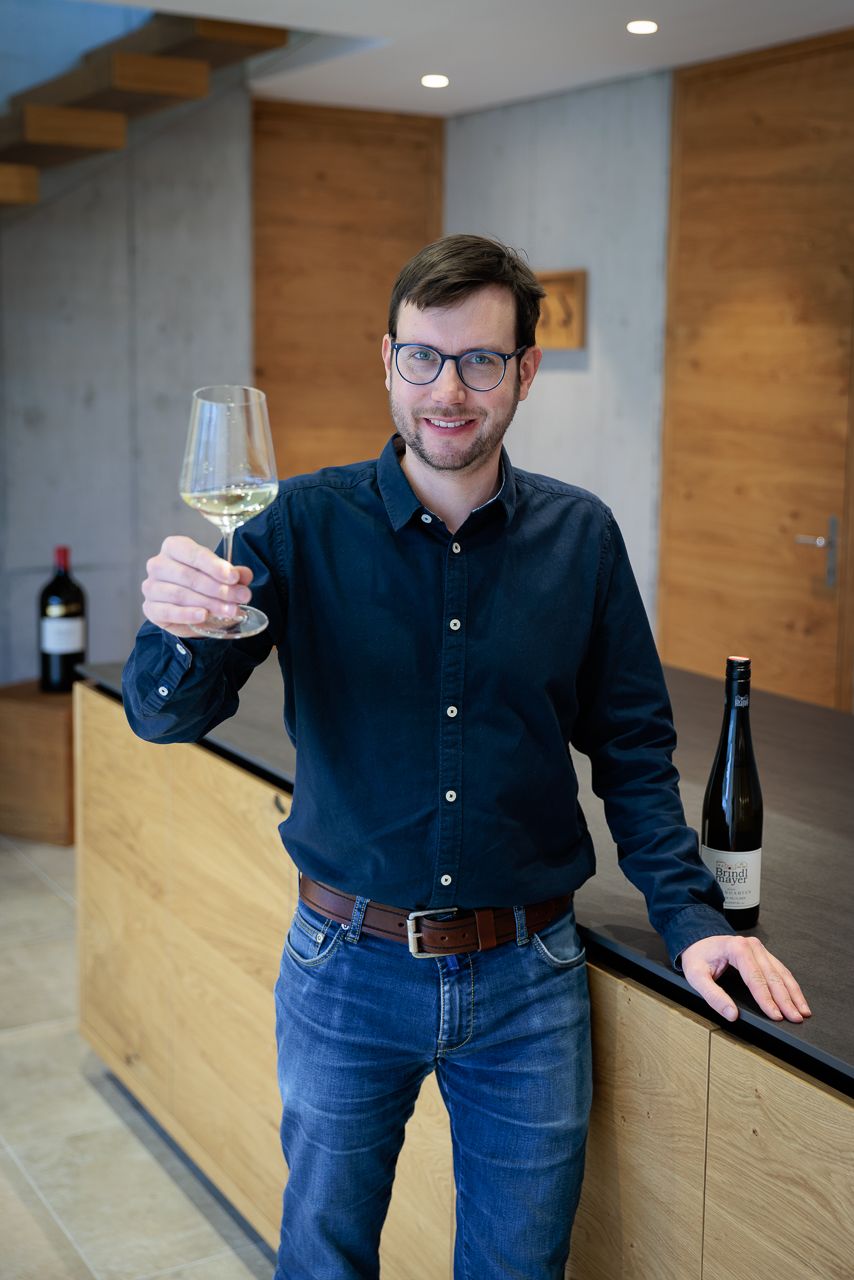 A man with glasses holds a glass of white wine in a modern room with wooden elements.