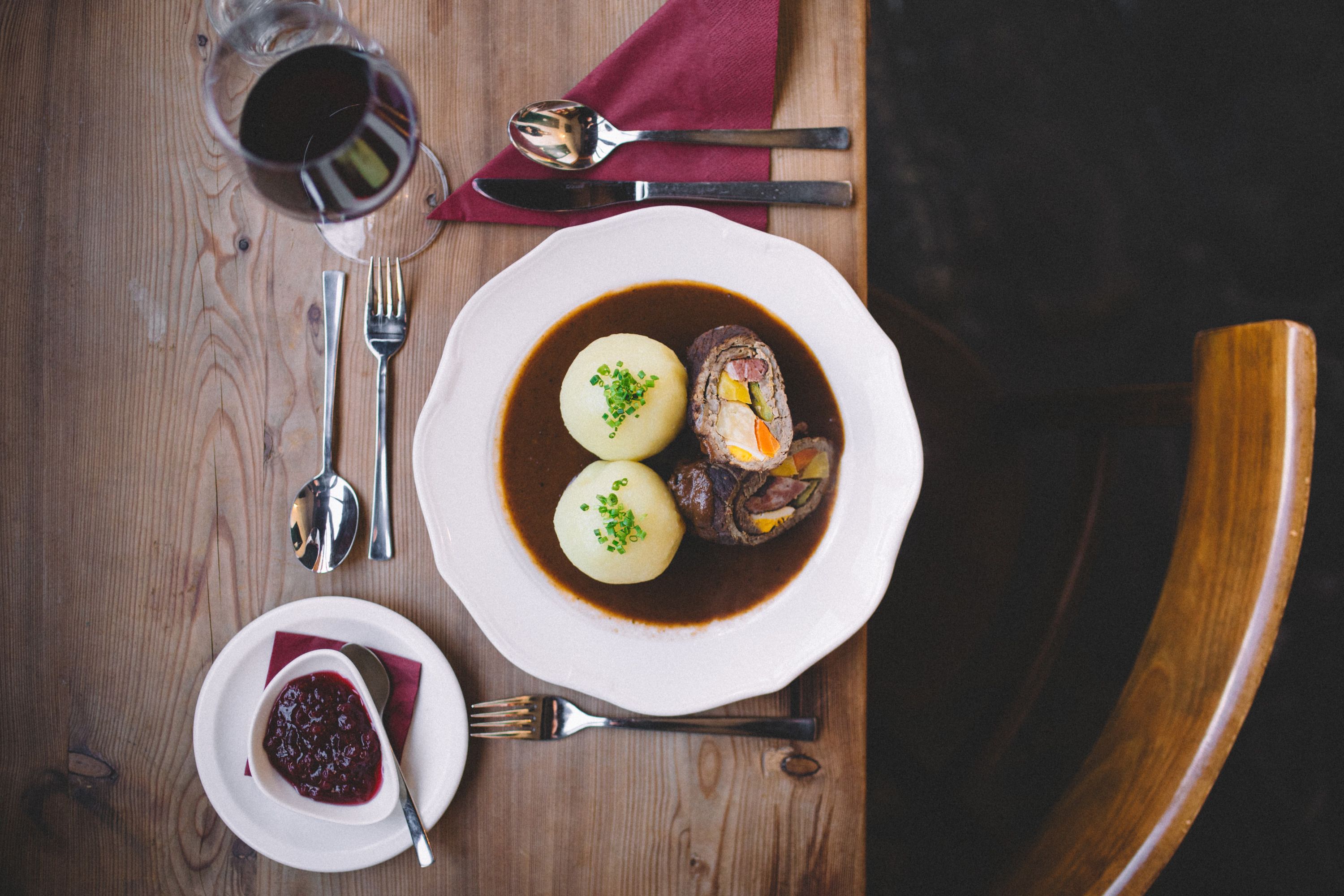 A plate with beef roulade and two potato dumplings in sauce, next to it a glass of red wine and cranberries on a wooden table.