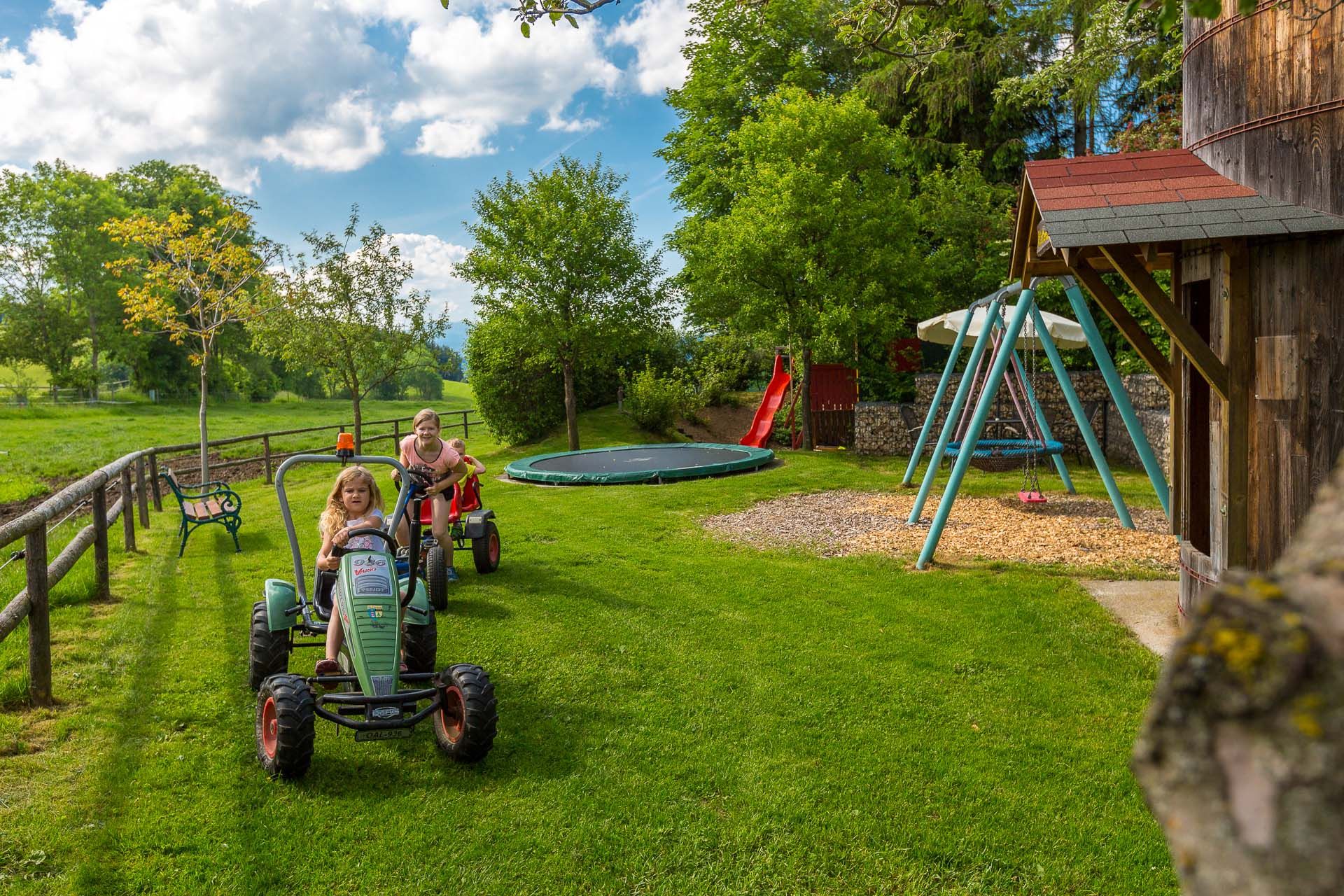 Two children ride toy tractors across a meadow. A swing, a slide and a trampoline can be seen in the background.