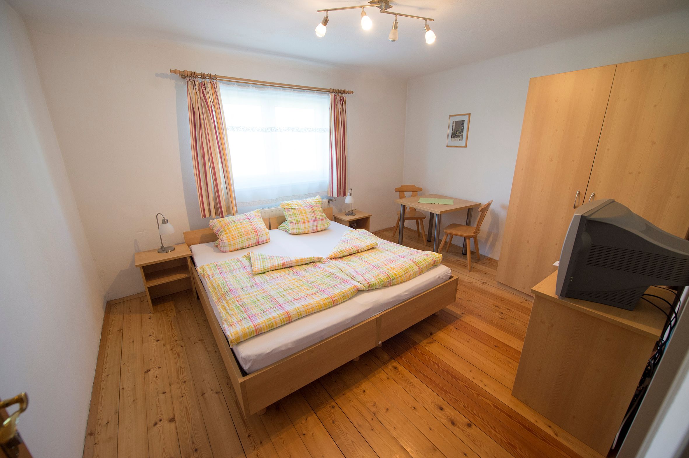 Room with double bed. Behind it a window, two chairs and a wardrobe with TV.