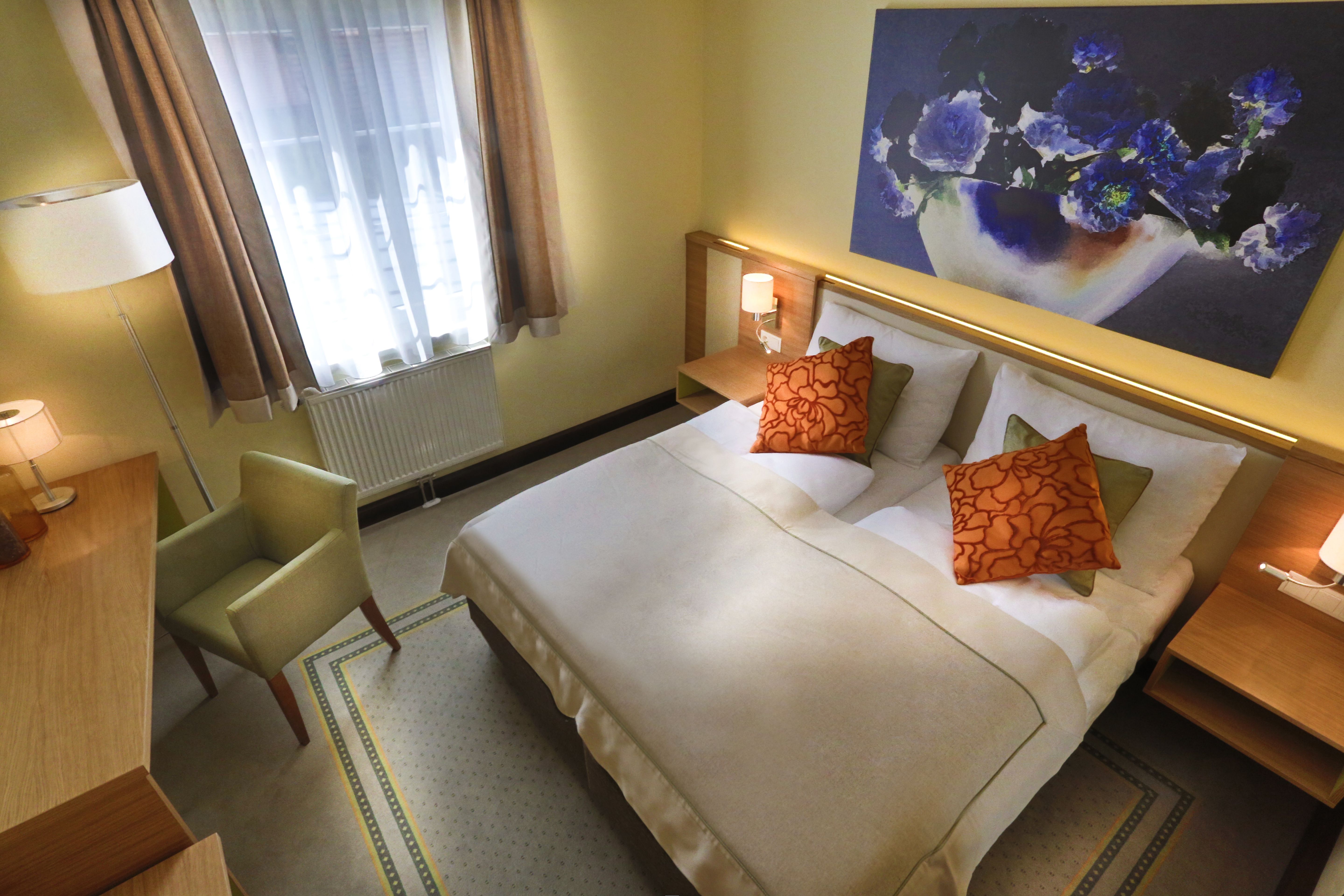 Cozy hotel room with a double bed, two orange pillows, a desk, an armchair and a large flower picture on the wall.