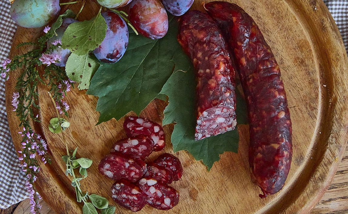 Kaminwurzn and plums on a wooden board decorated with green leaves and herbs.