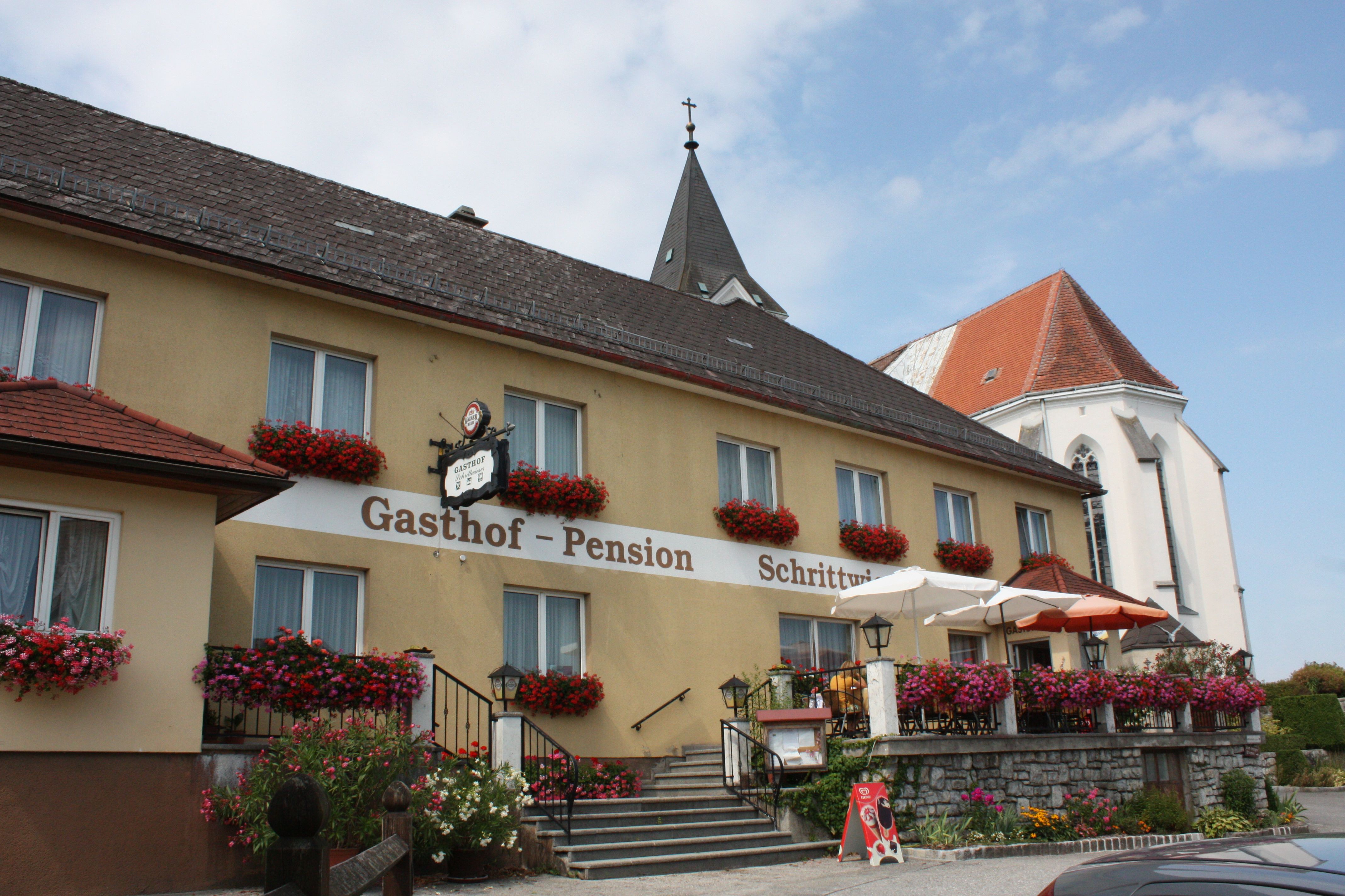 A traditional inn with a flower-decorated façade and terrace, next to a church with a pointed tower.