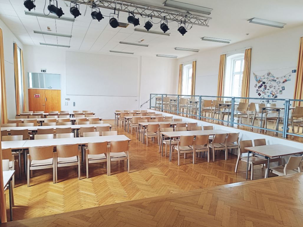 An empty hall with a wooden floor, tables and chairs, a stage and lighting on the ceiling.
