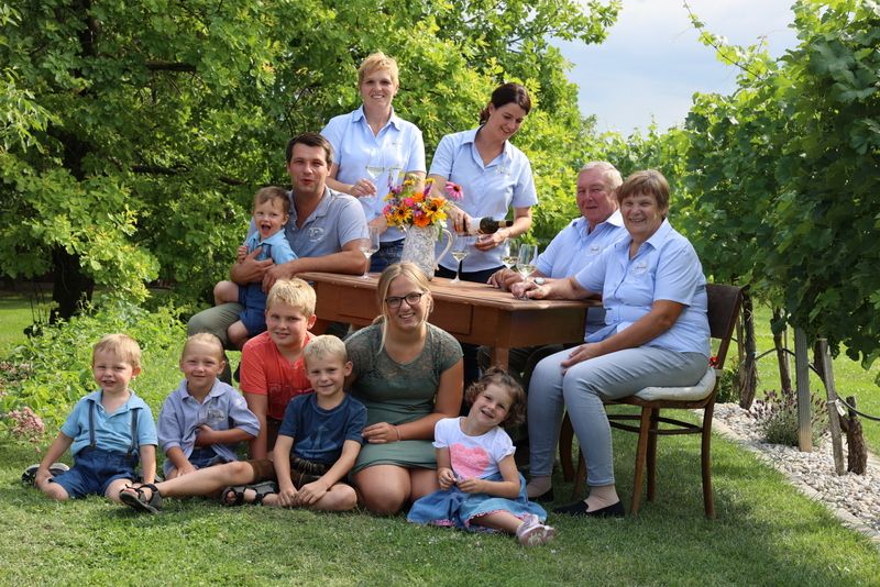 A family sits and stands around an outdoor table, surrounded by trees and grass.