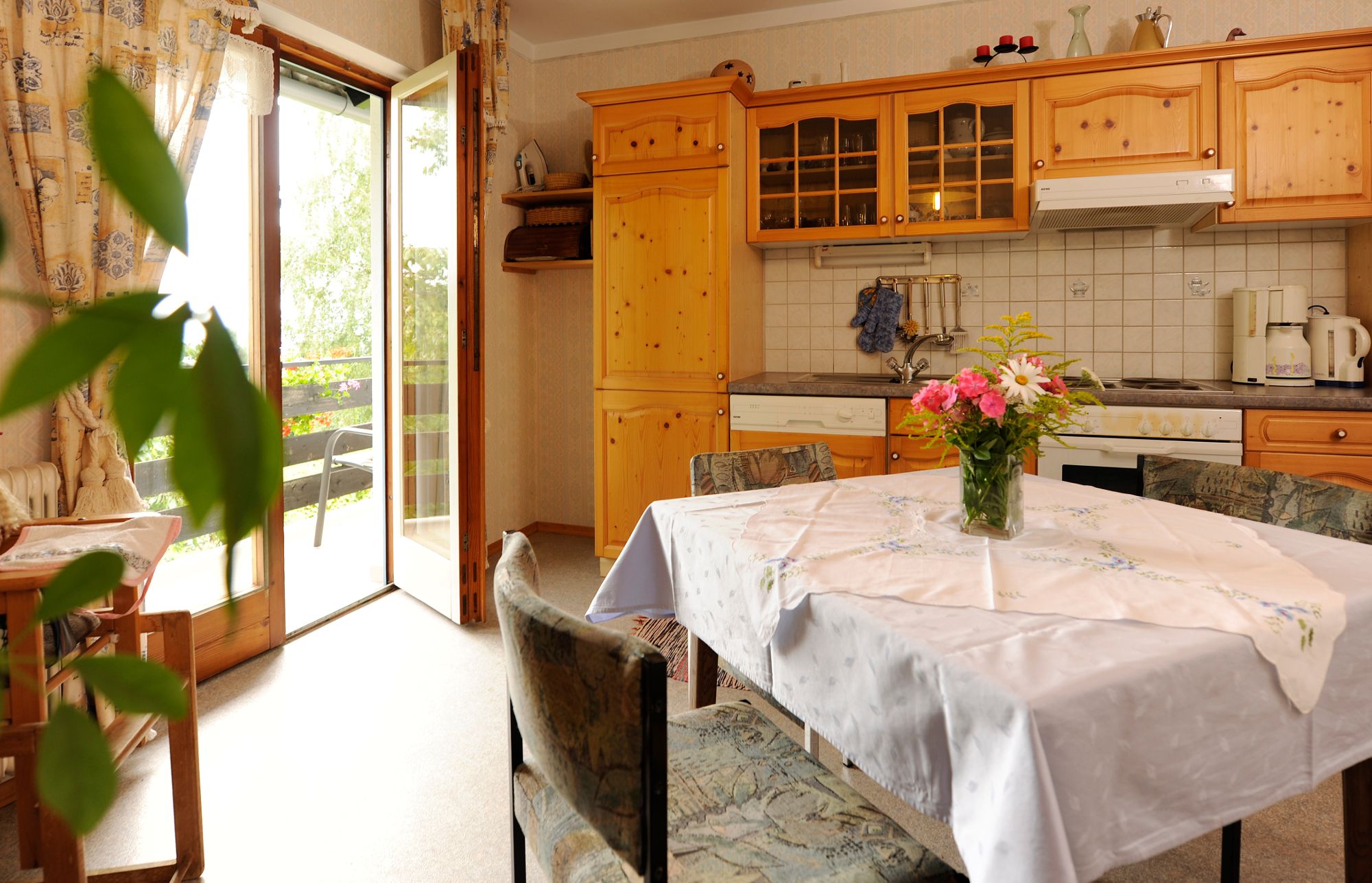 Cozy kitchen with wooden furniture, dining table and bouquet of flowers. Open balcony door with a view of the greenery.