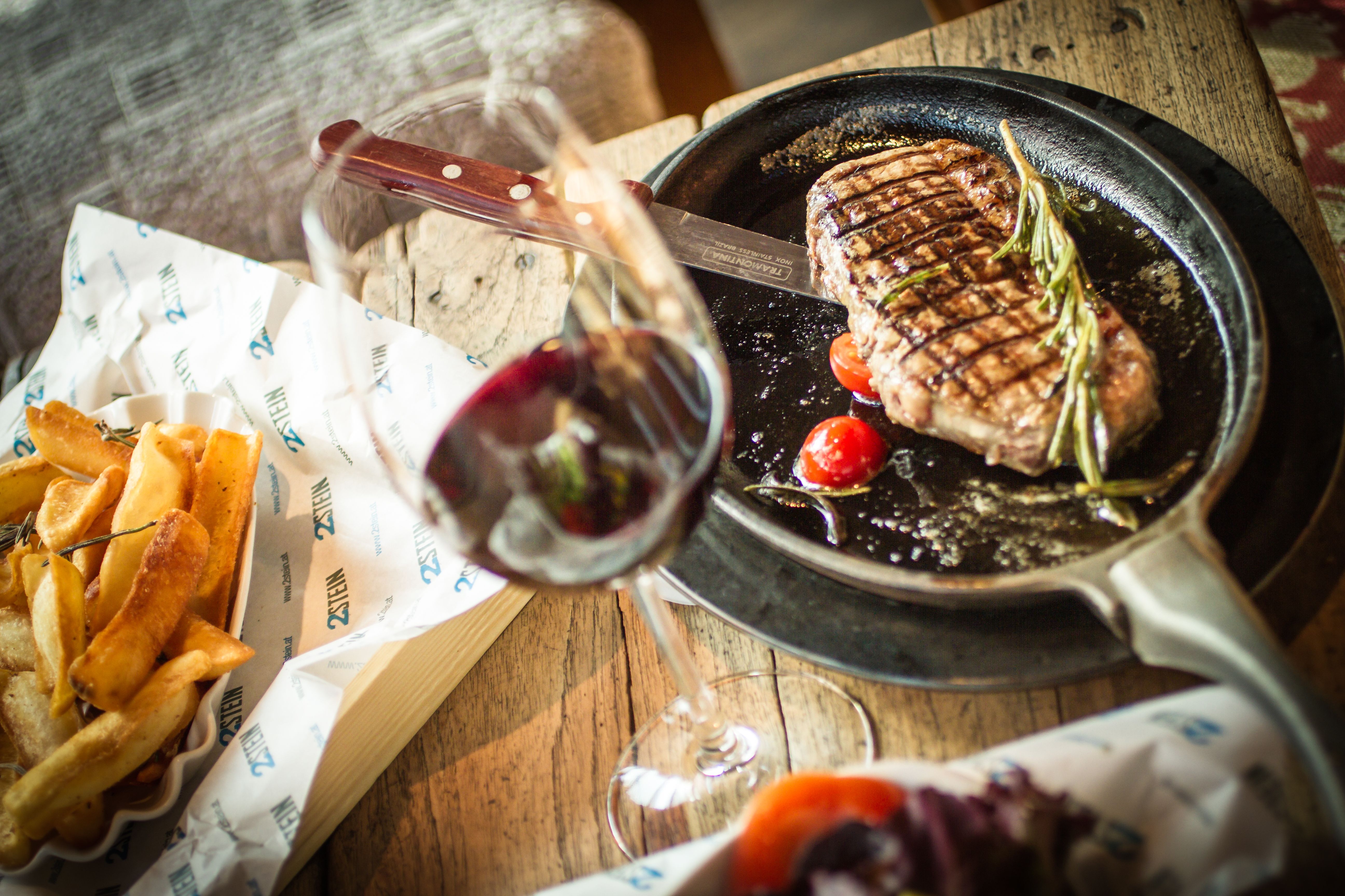 A steak in a pan with tomatoes and rosemary, French fries and a glass of red wine on a wooden table next to it.