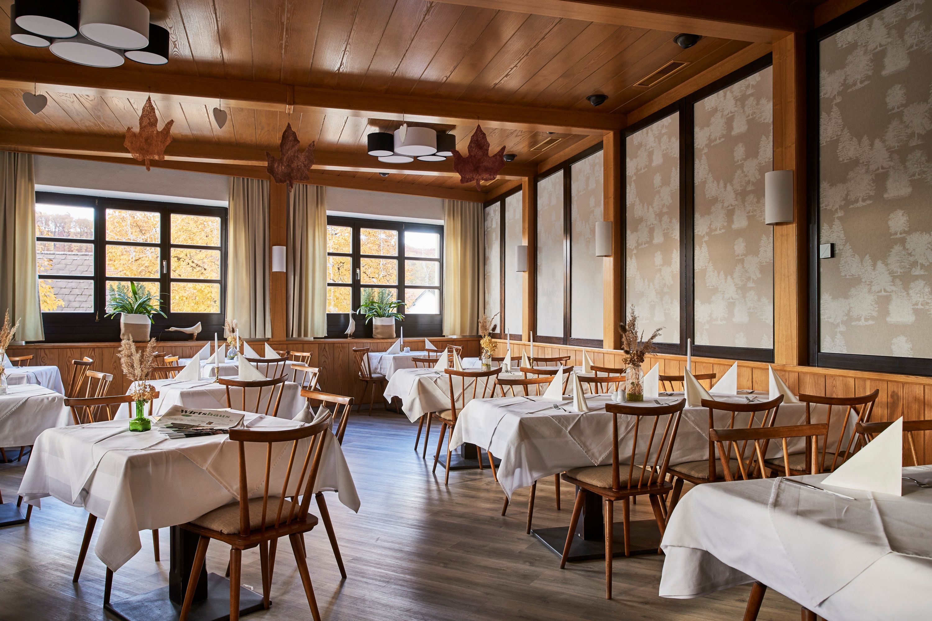 An elegant restaurant with wooden furniture, white tablecloths and large windows showing autumnal trees.