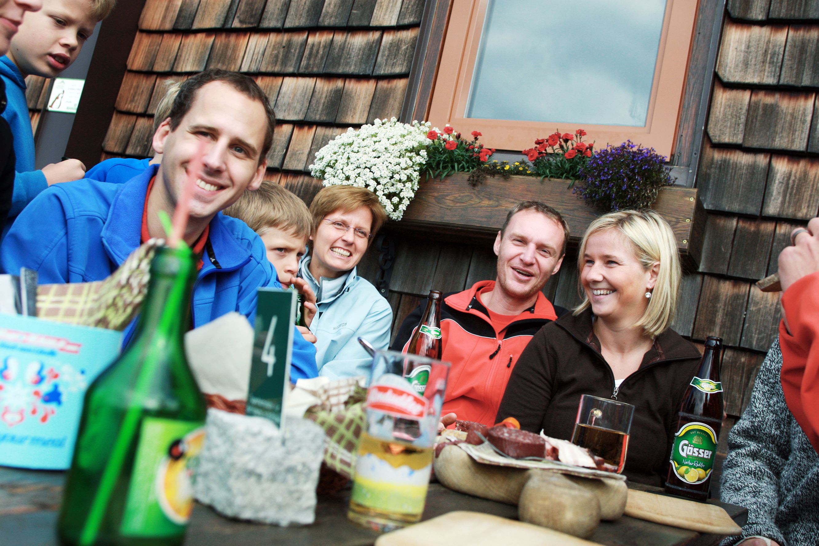 Group of people sitting laughing at a table in front of a wooden hut with flowers.