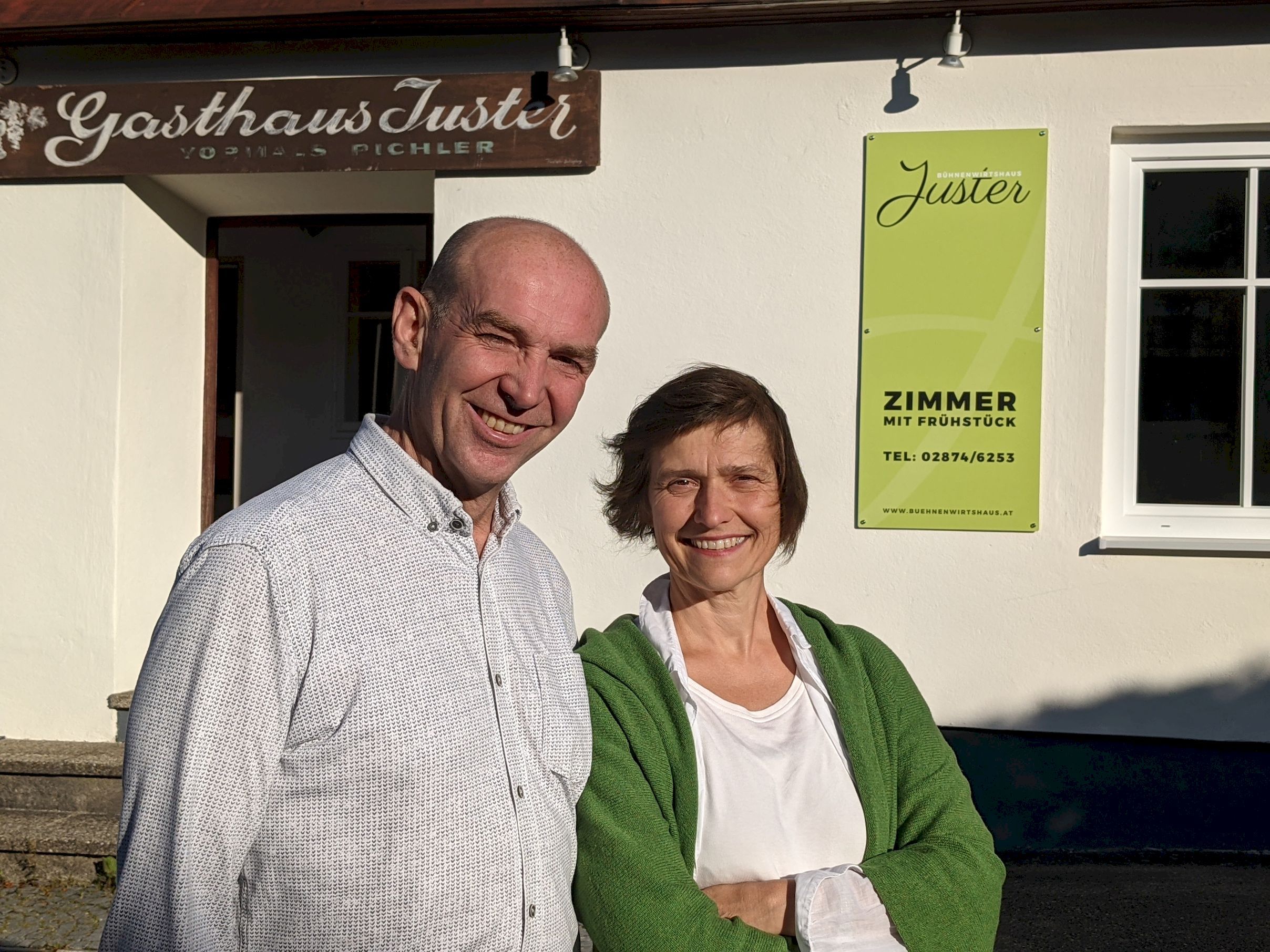 A man and a woman stand smiling in front of the Jüster inn. A sign reading 'Room with breakfast' can be seen in the background.