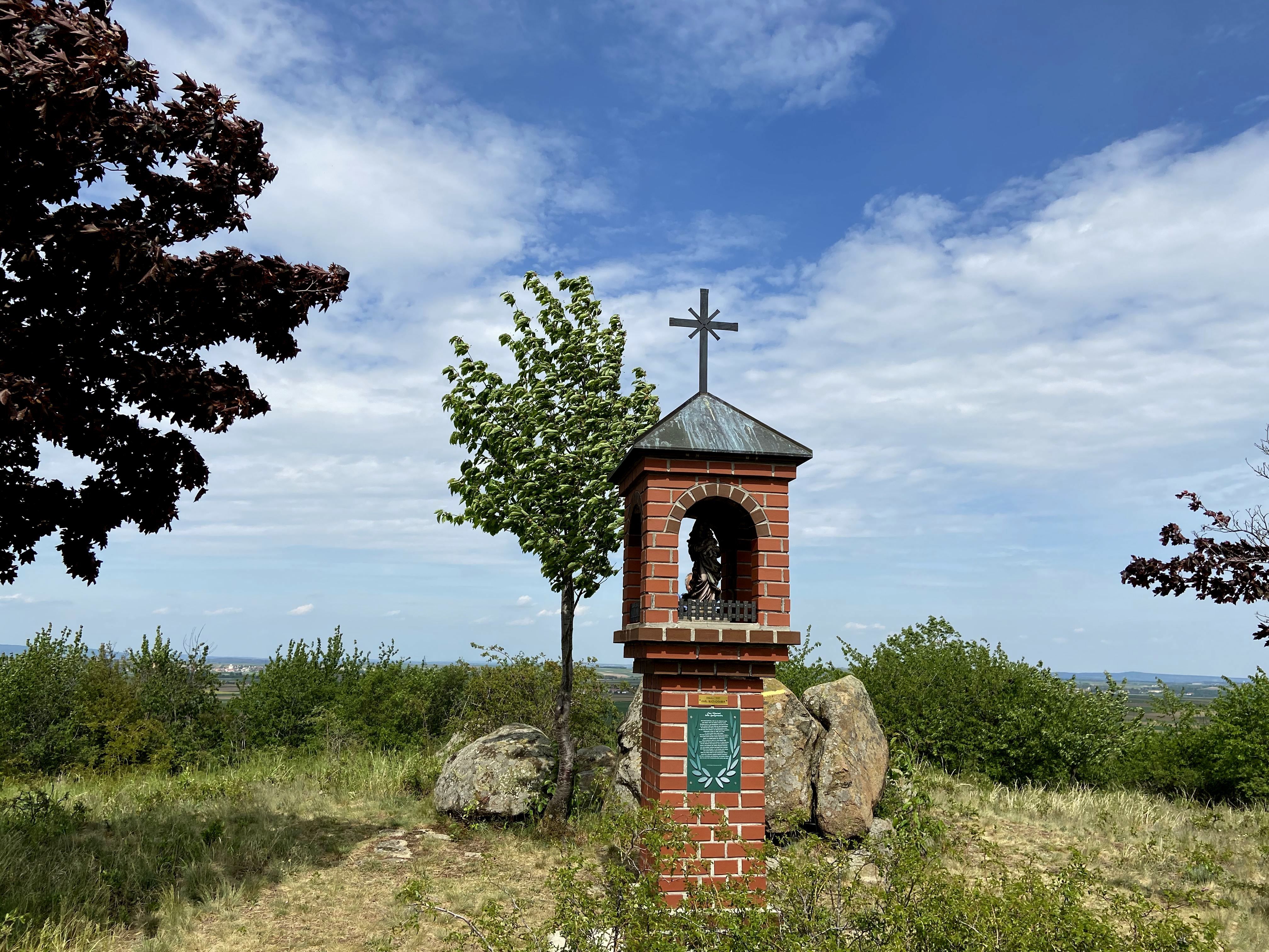 A brick wayside shrine with a cross on a hill, surrounded by trees and bushes, under a blue sky.