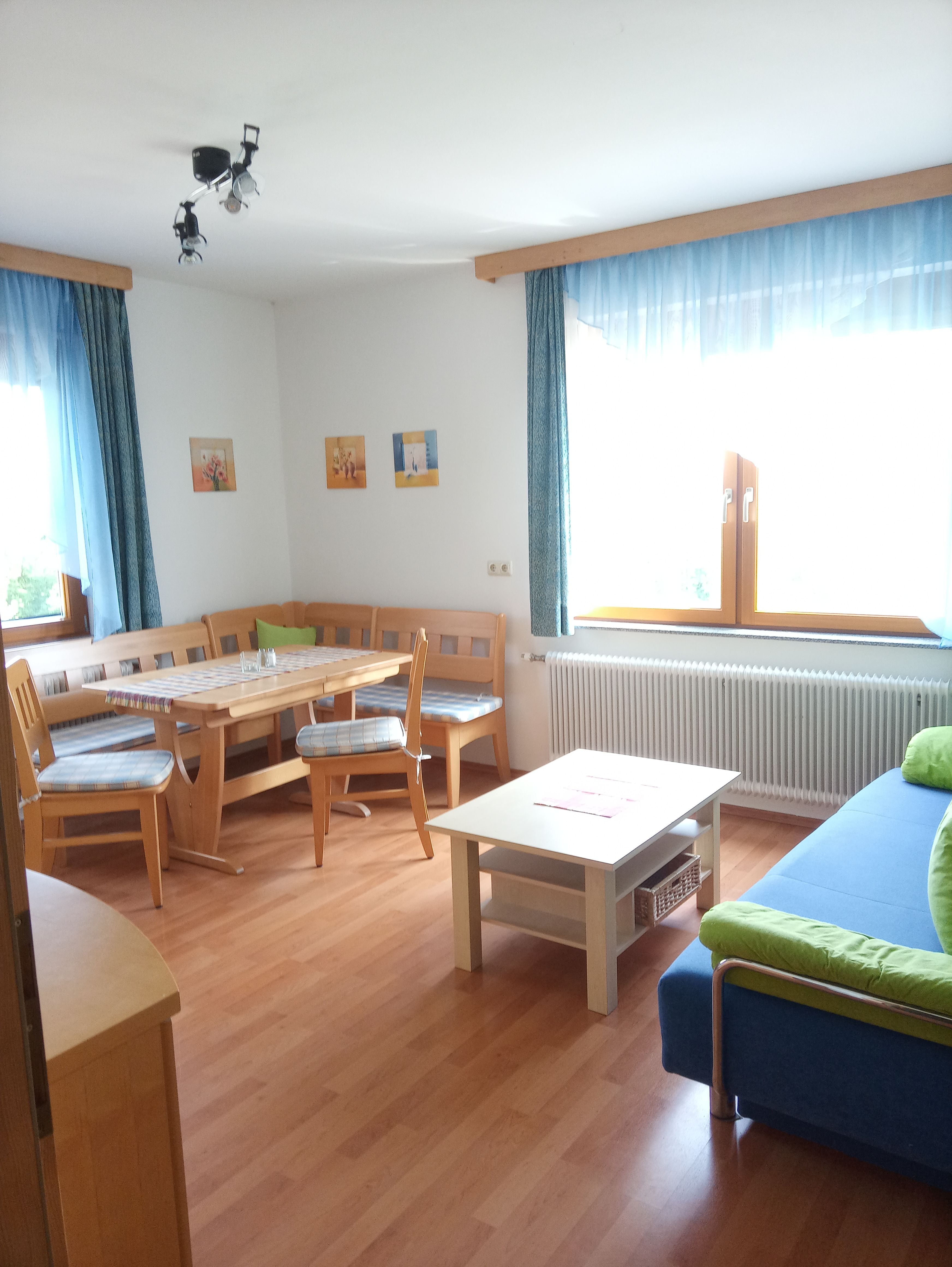 Bright living room with wooden furniture, corner bench, table, sofa and large windows with blue curtains.