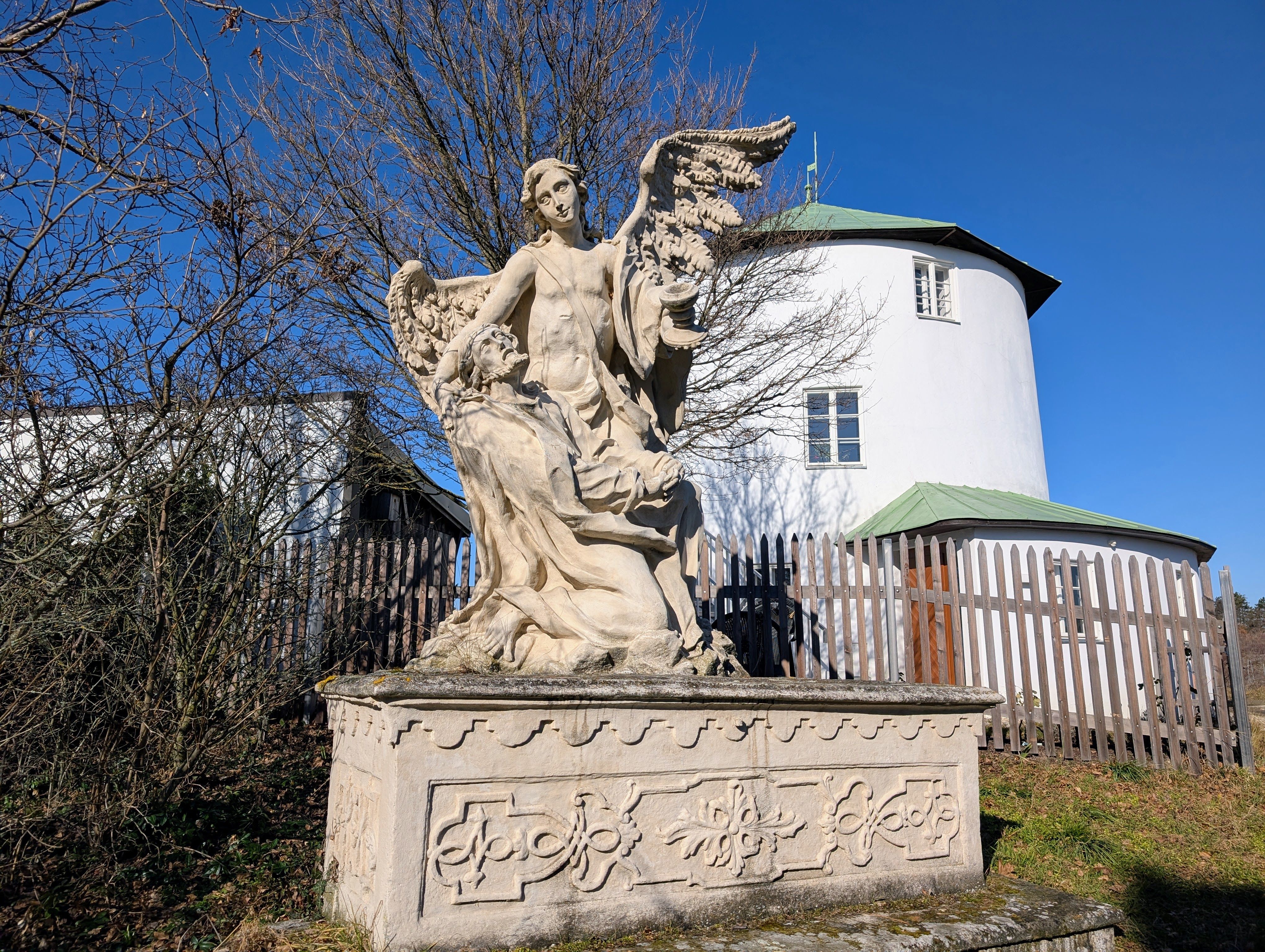 Stone statue of an angel with wings and a kneeling figure in front of a round, white building with a green roof.