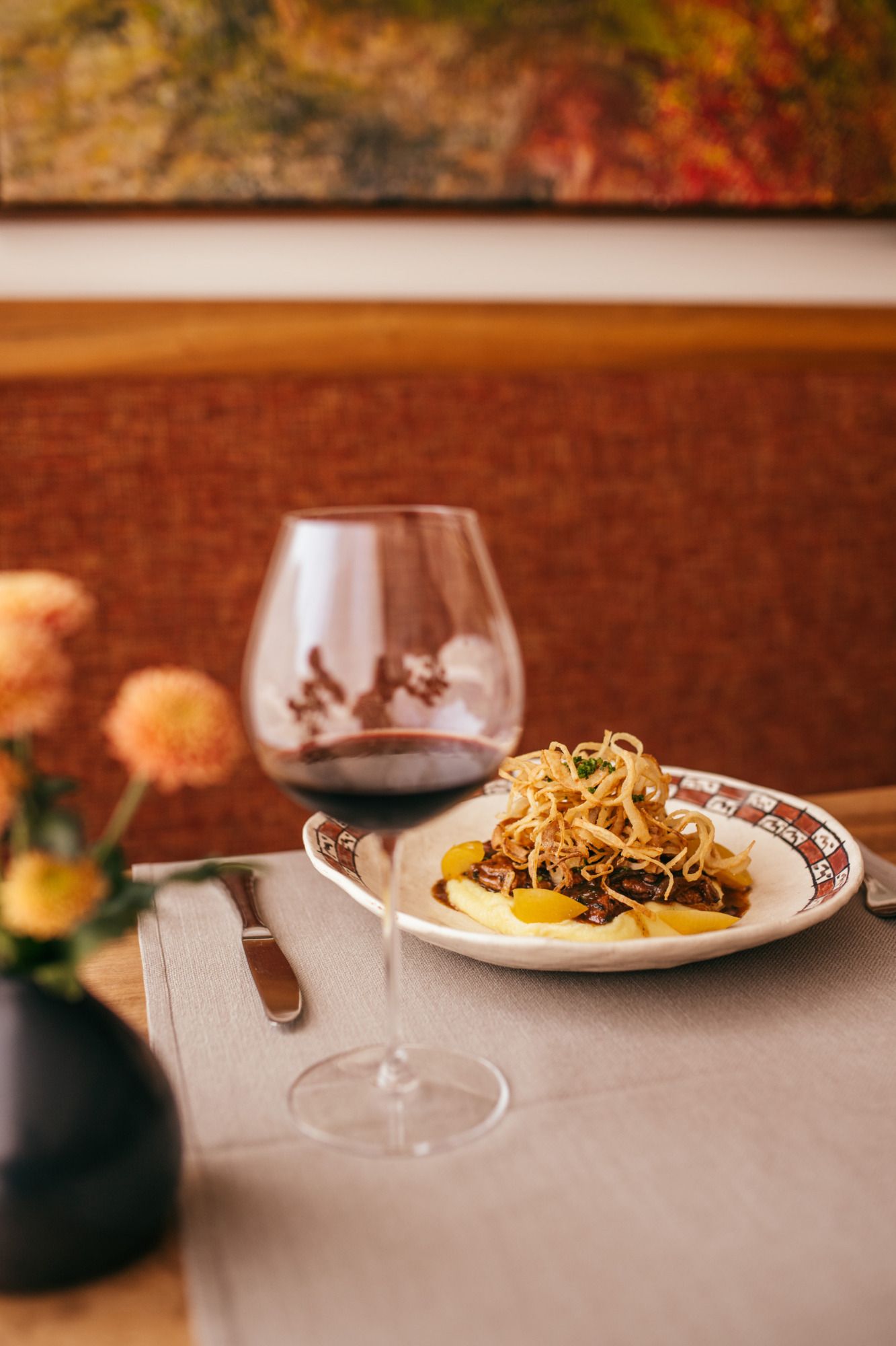 A plate of glazed veal liver on mashed potatoes, garnished with fried onions, next to a glass of red wine on a table.