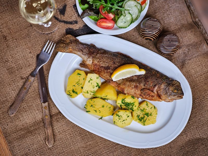 A plate with fried trout, potatoes and a slice of lemon, next to a salad and a glass of white wine.