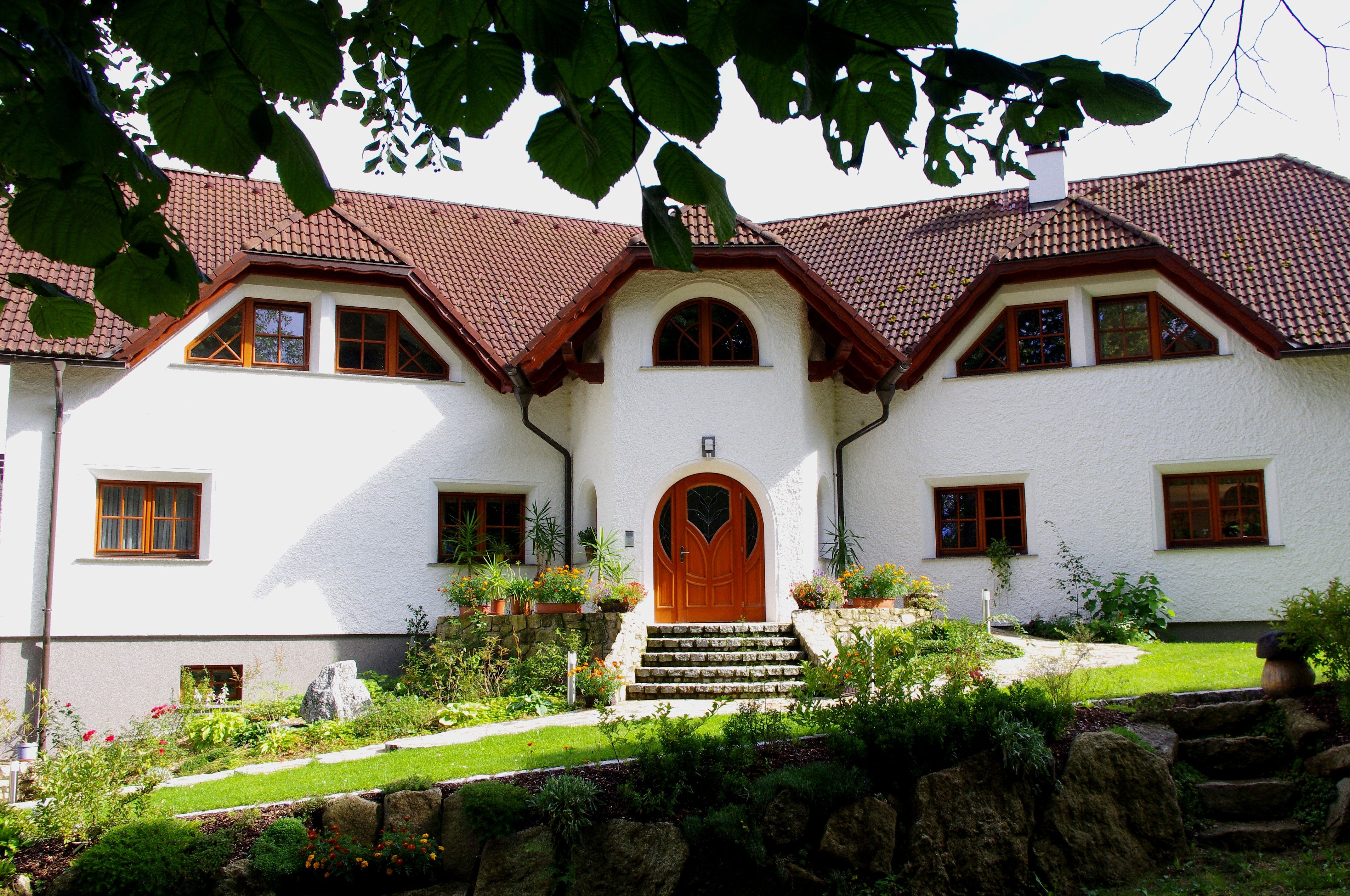 A large, white house with a red tiled roof and a well-tended garden surrounded by trees.
