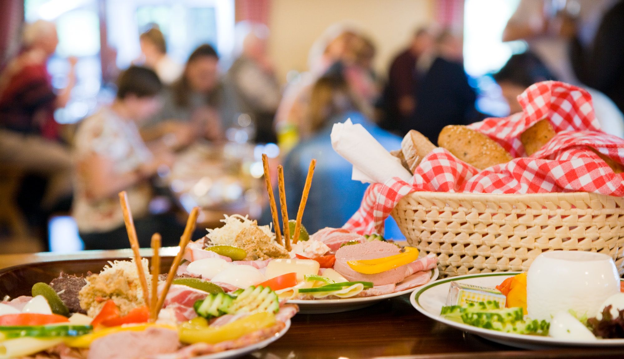 A table with traditional Austrian dishes, including cold cuts, cheese and bread in a basket with a red and white checked cloth.