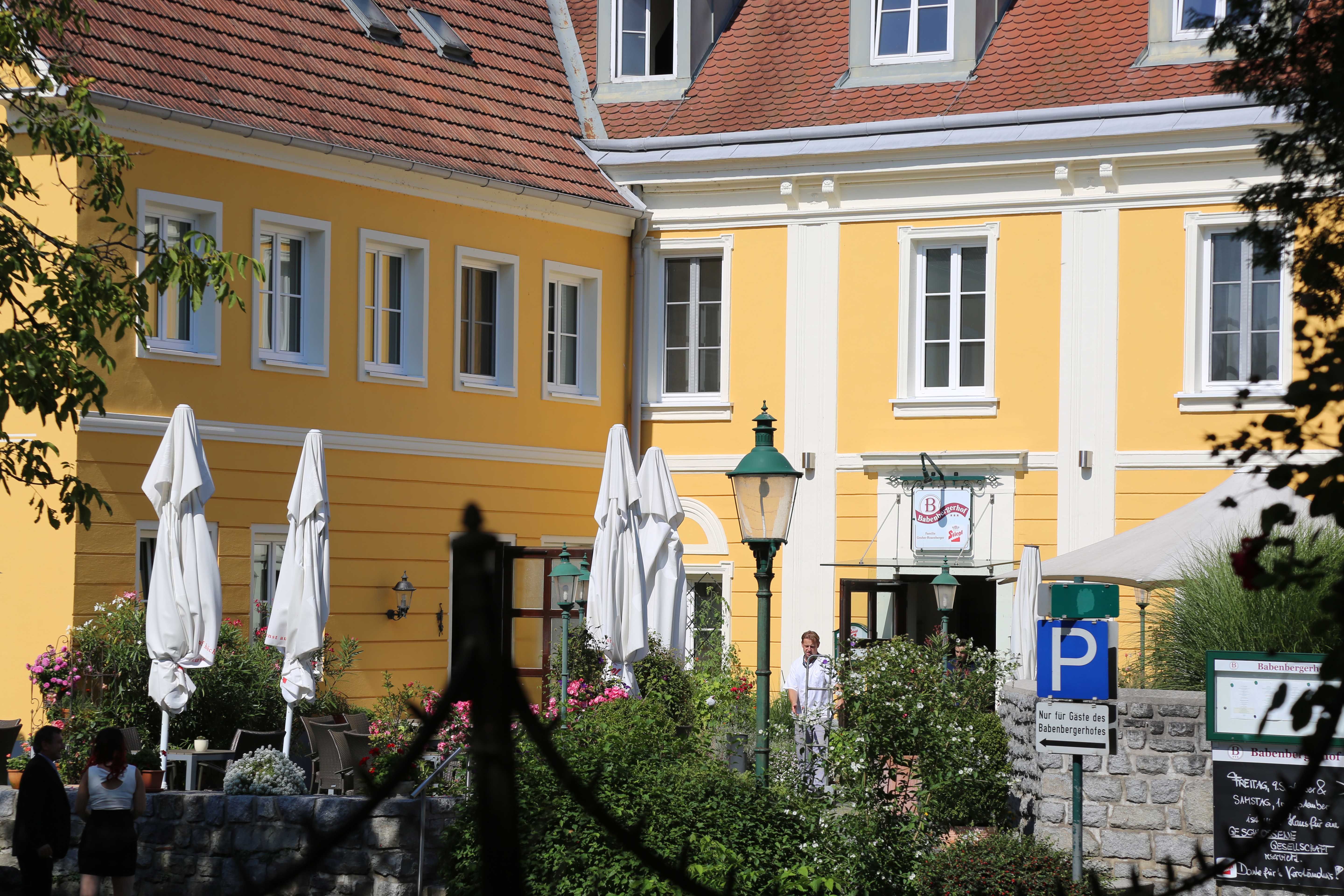 Exterior view of a yellow building with red roof tiles and white window frames, surrounded by plants and parasols.