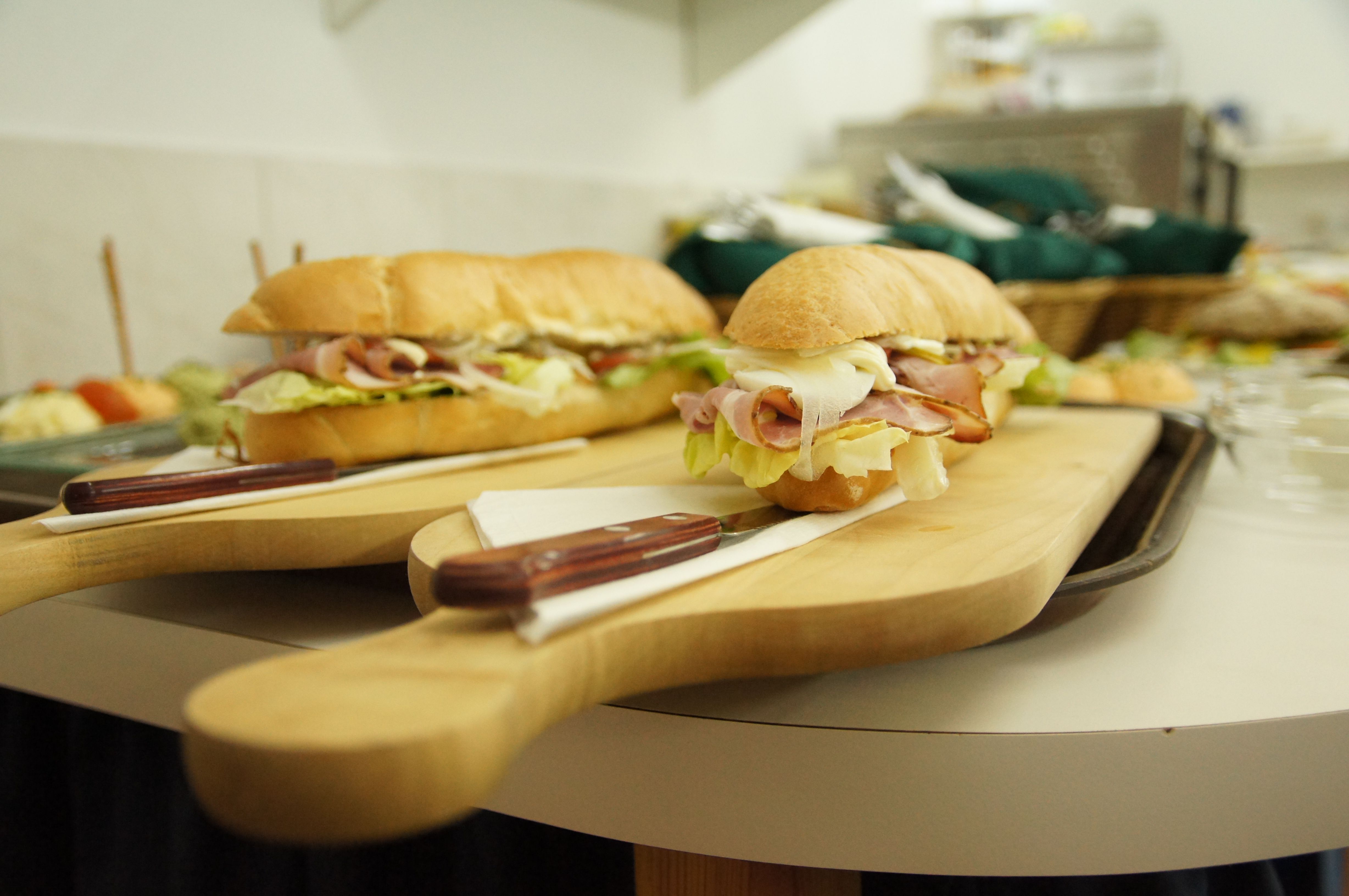 Two filled baguettes on a wooden board with a knife, other dishes in the background.