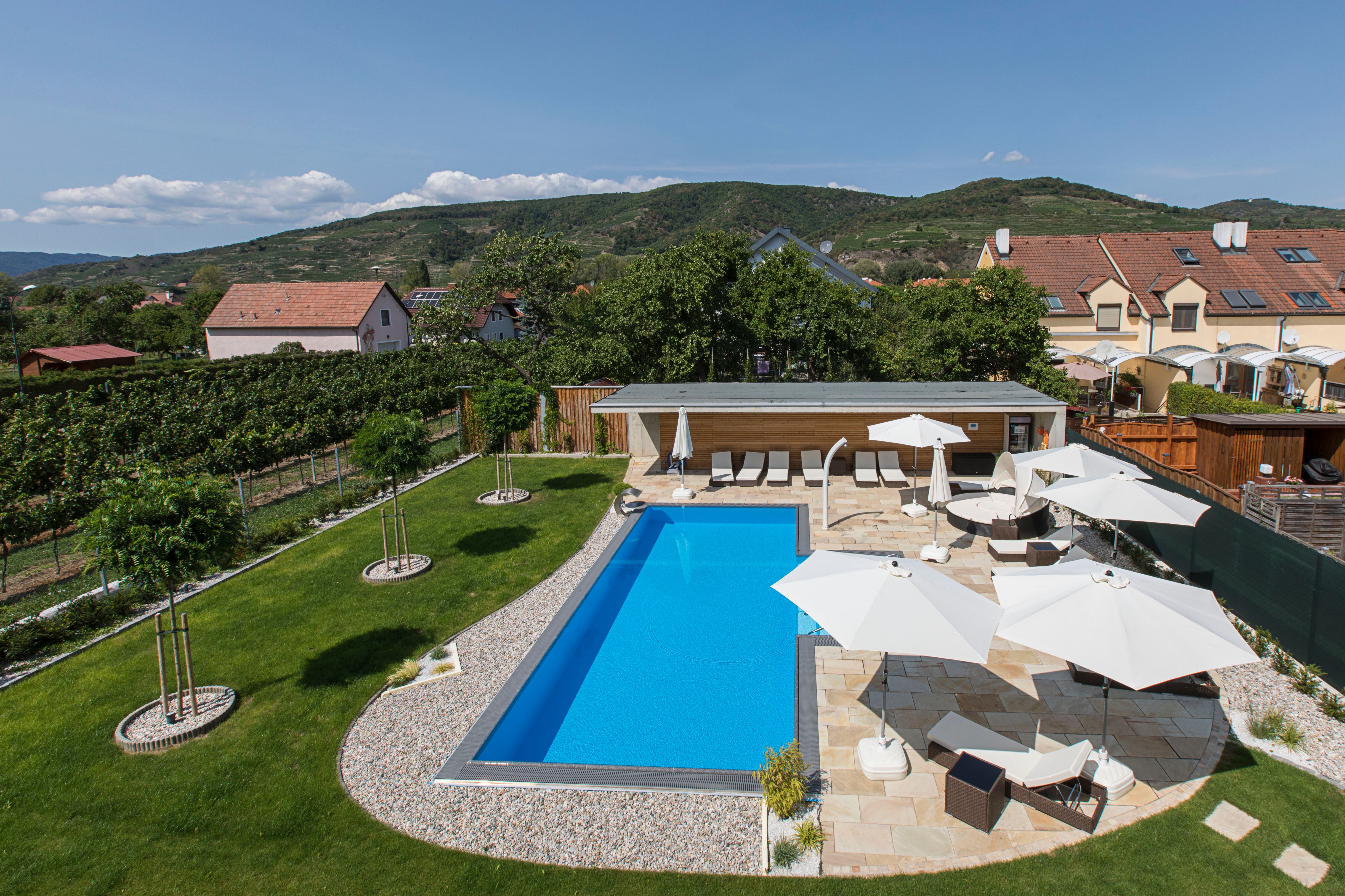 A modern pool with sun loungers and parasols in a well-tended garden surrounded by hills and houses.