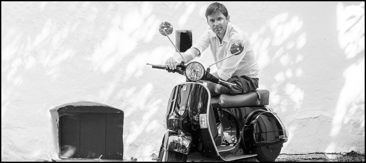 Person on a Vespa in front of a white wall.