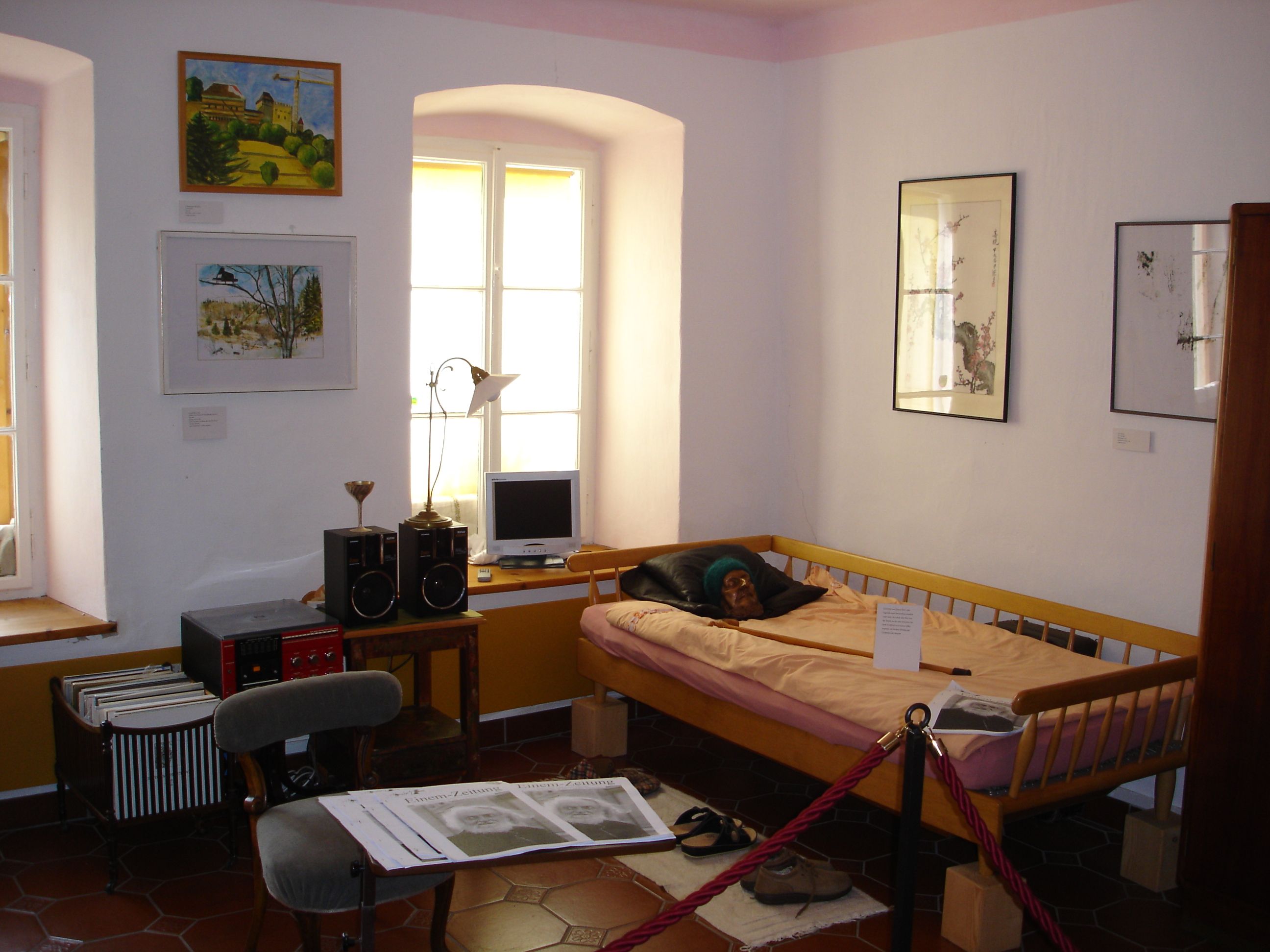Interior view of a museum room with a bed, pictures on the wall and a small desk with a computer.