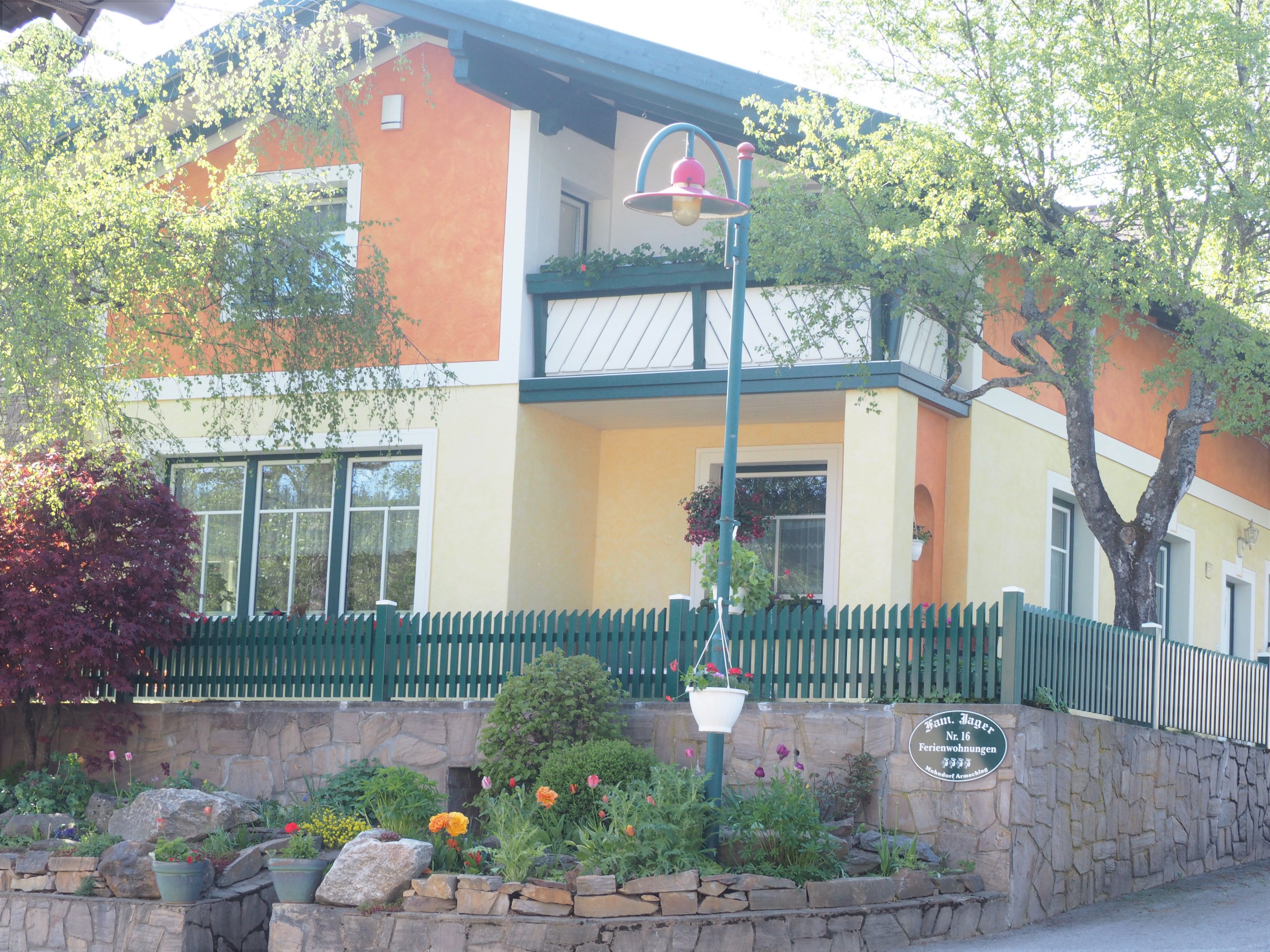 A two-storey house with an orange and yellow façade, green fence and garden.