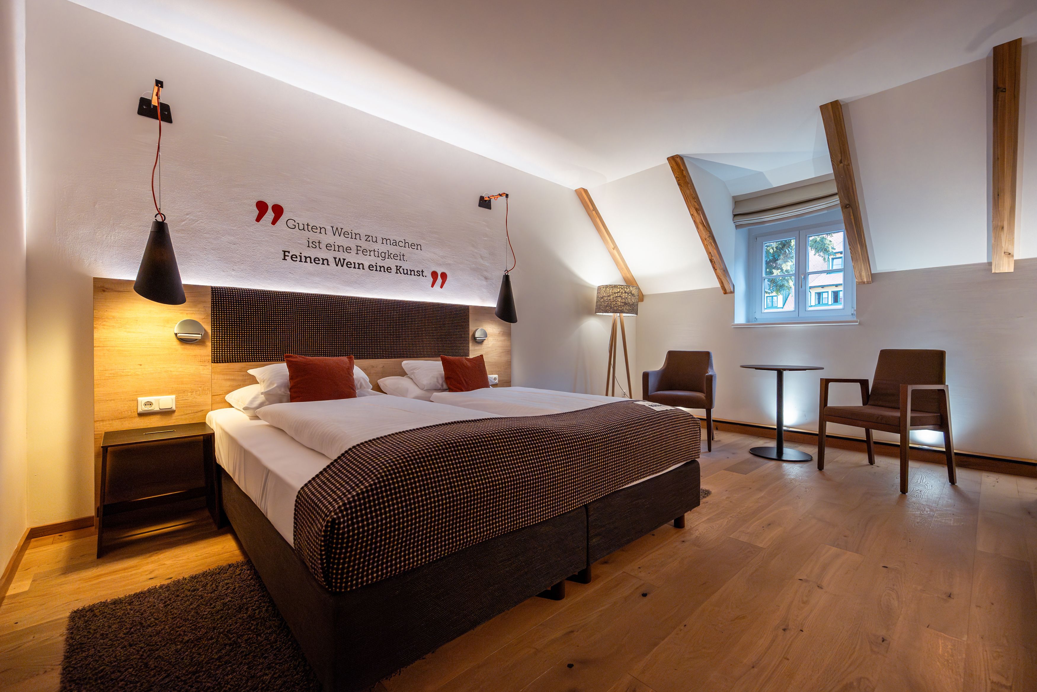 A stylishly furnished hotel room with a double bed, two armchairs and a small table. There is a quote about wine on the wall.