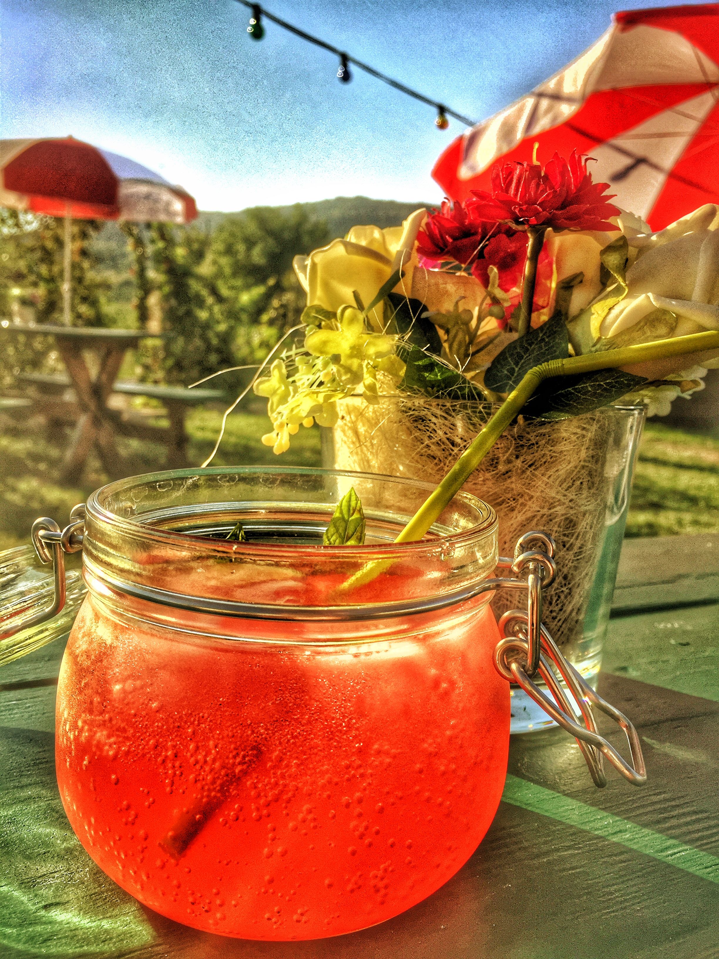 A red drink in a glass with a swing top, next to it a bouquet of flowers on an outdoor table.