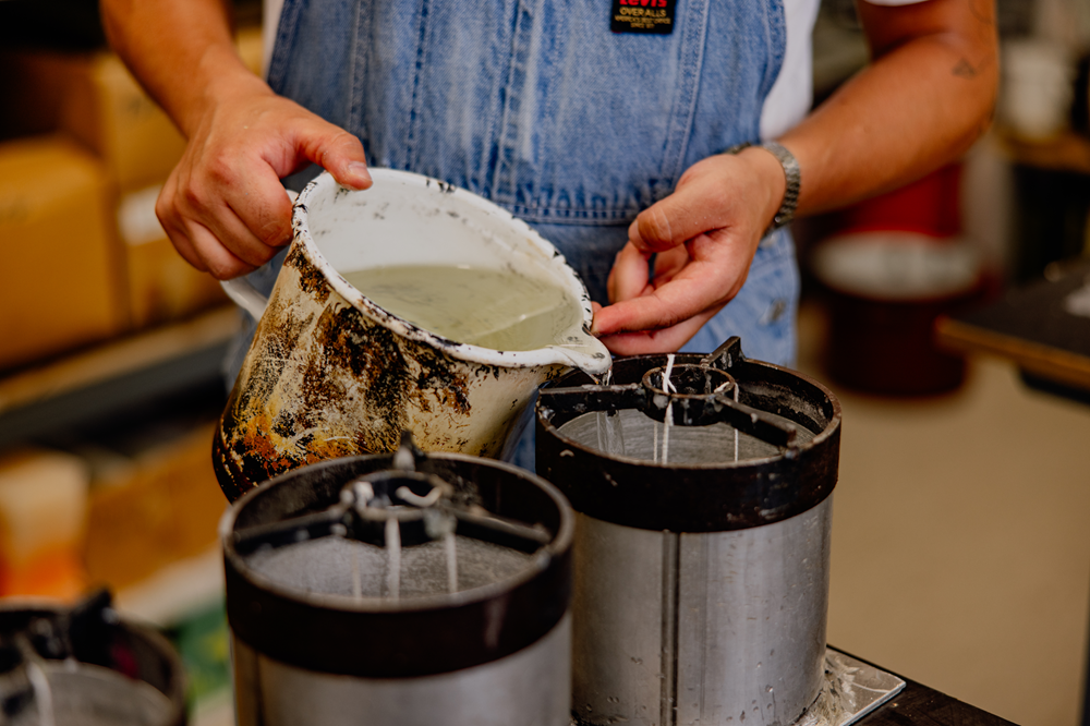 Person pours liquid wax from a kettle into a candle mold.