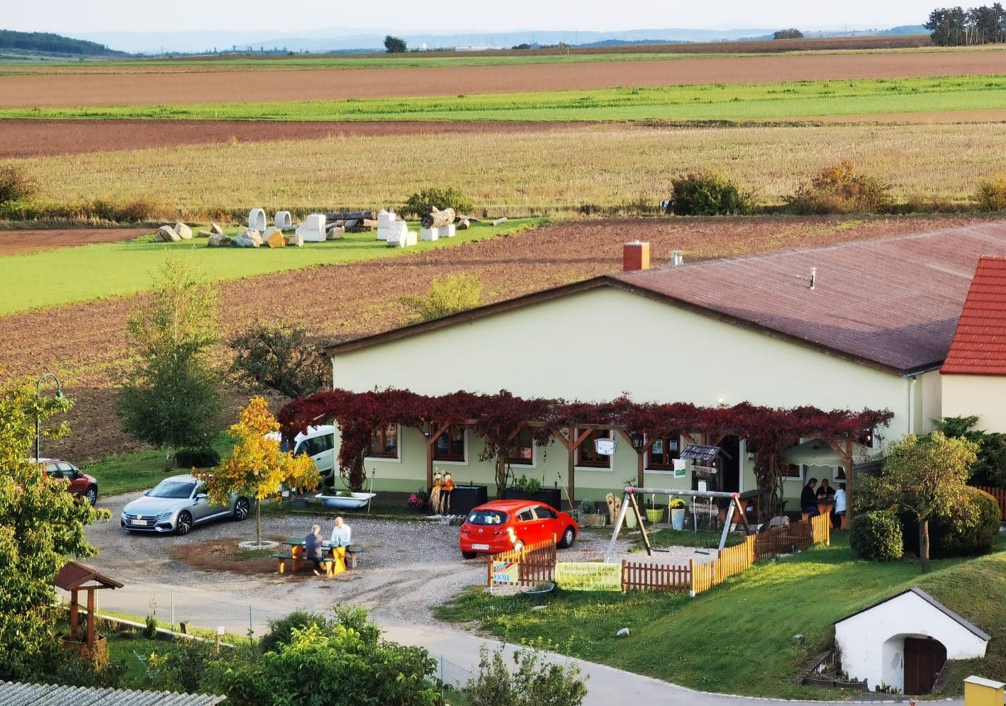 A rural building covered in vines, surrounded by fields and cars. People are sitting at a table outside.
