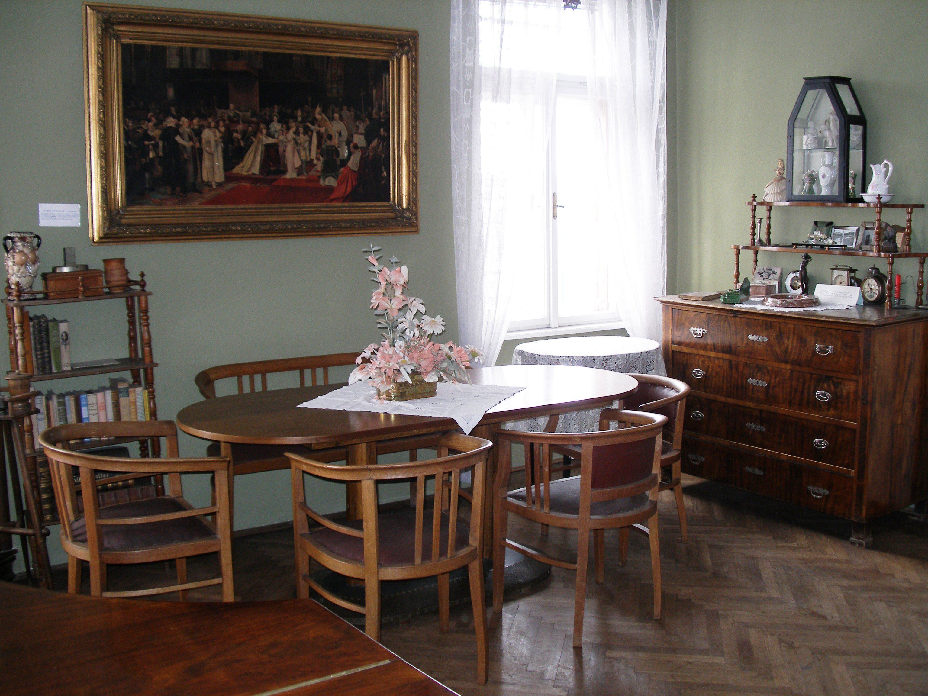 A traditionally furnished living room with wooden furniture, a round table, chairs, a chest of drawers and a large painting on the wall.
