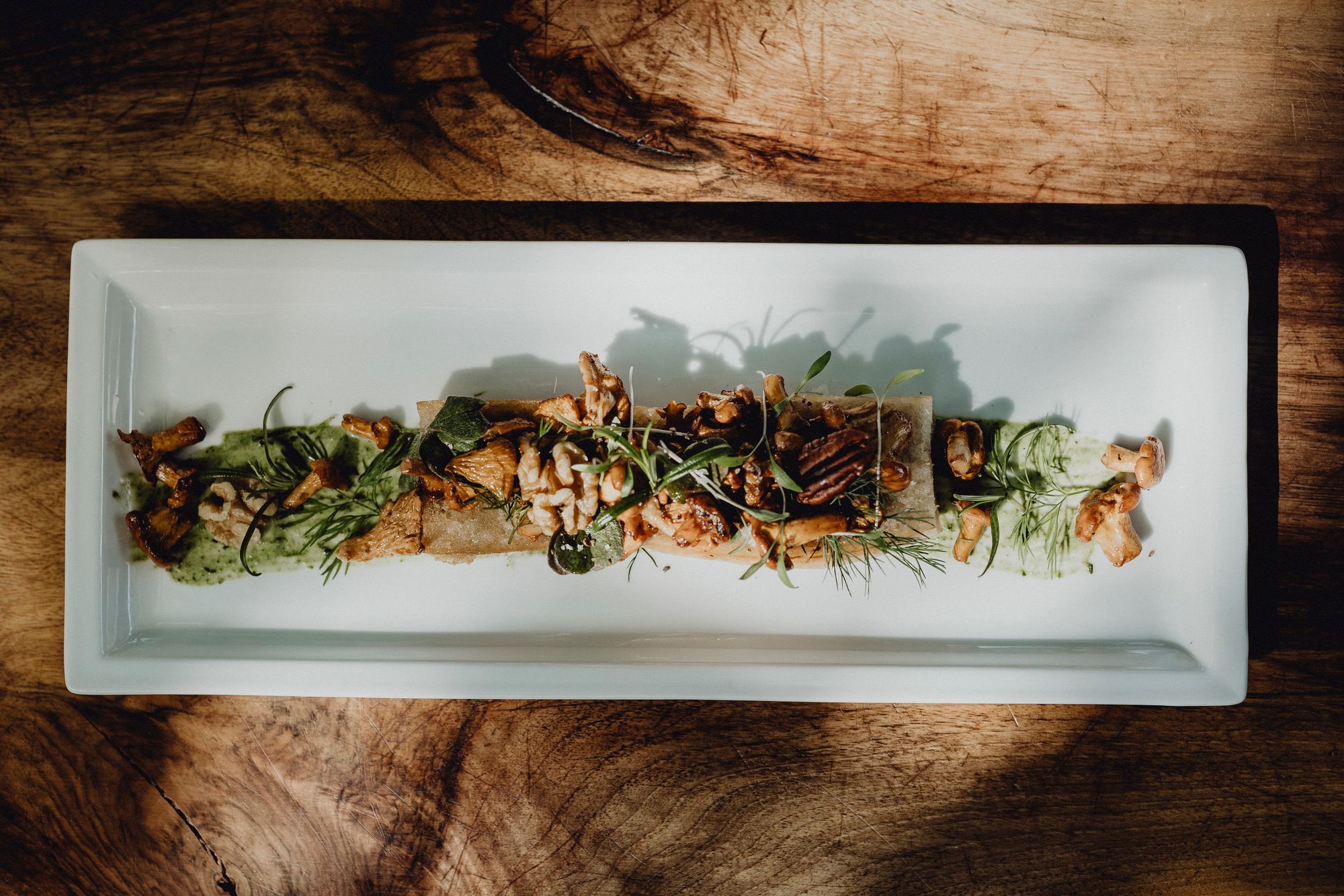 A rectangular plate with sautéed chanterelles, pecan nuts, wild herbs and root bread, artfully arranged on a wooden table.