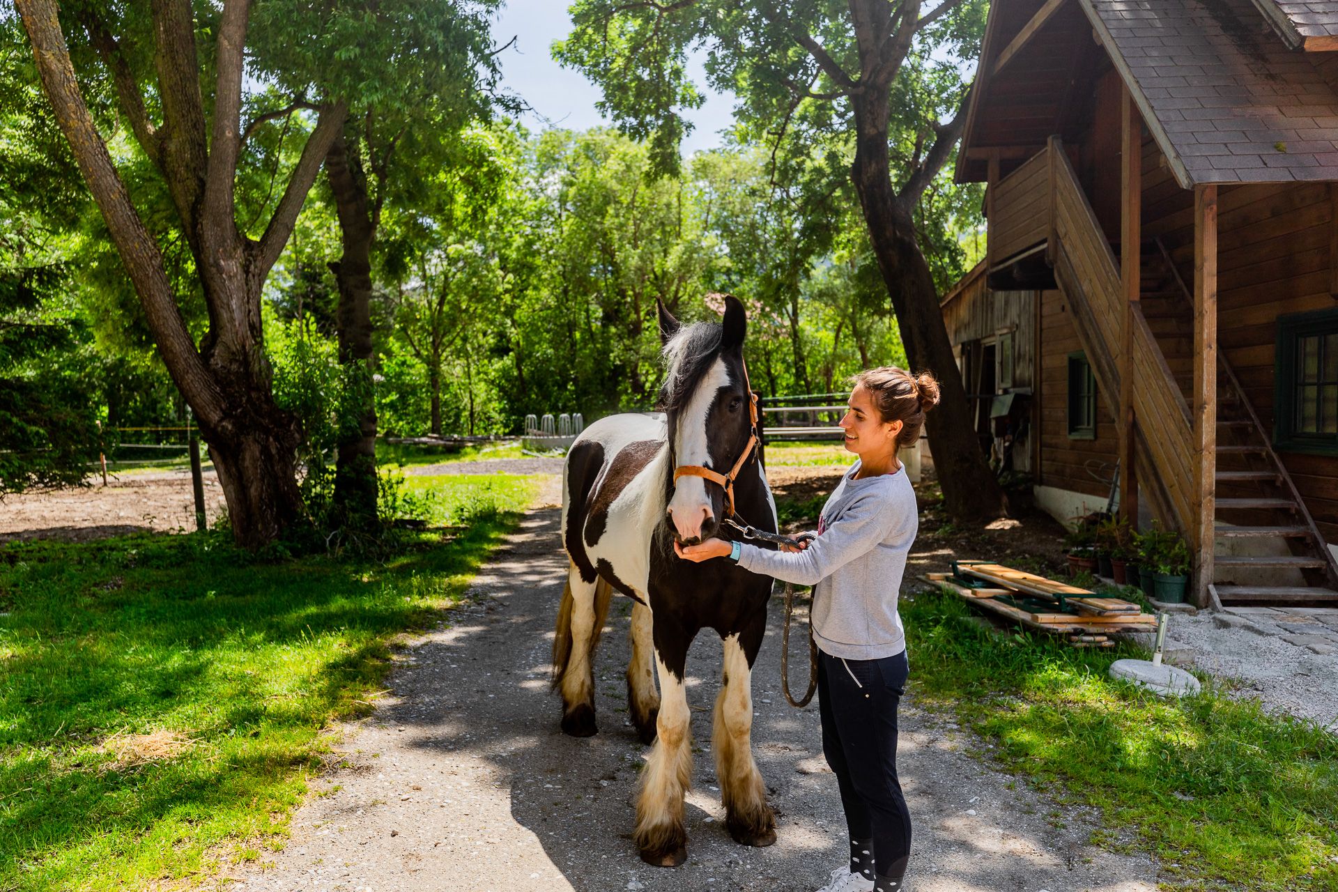 A woman stands next to a black and white horse in a courtyard with trees and a wooden building in the background.