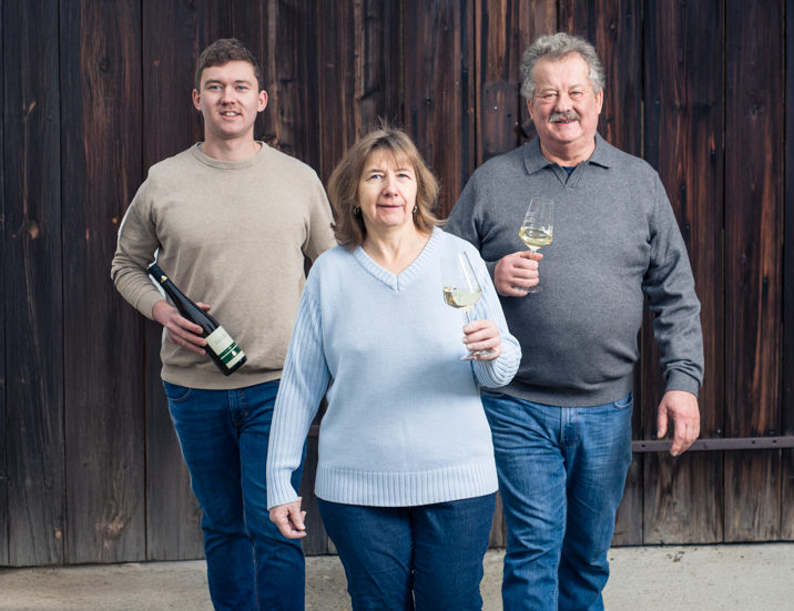 Three people are standing in front of a wooden façade. Two men and a woman are each holding a wine glass, one person is also holding a wine bottle.
