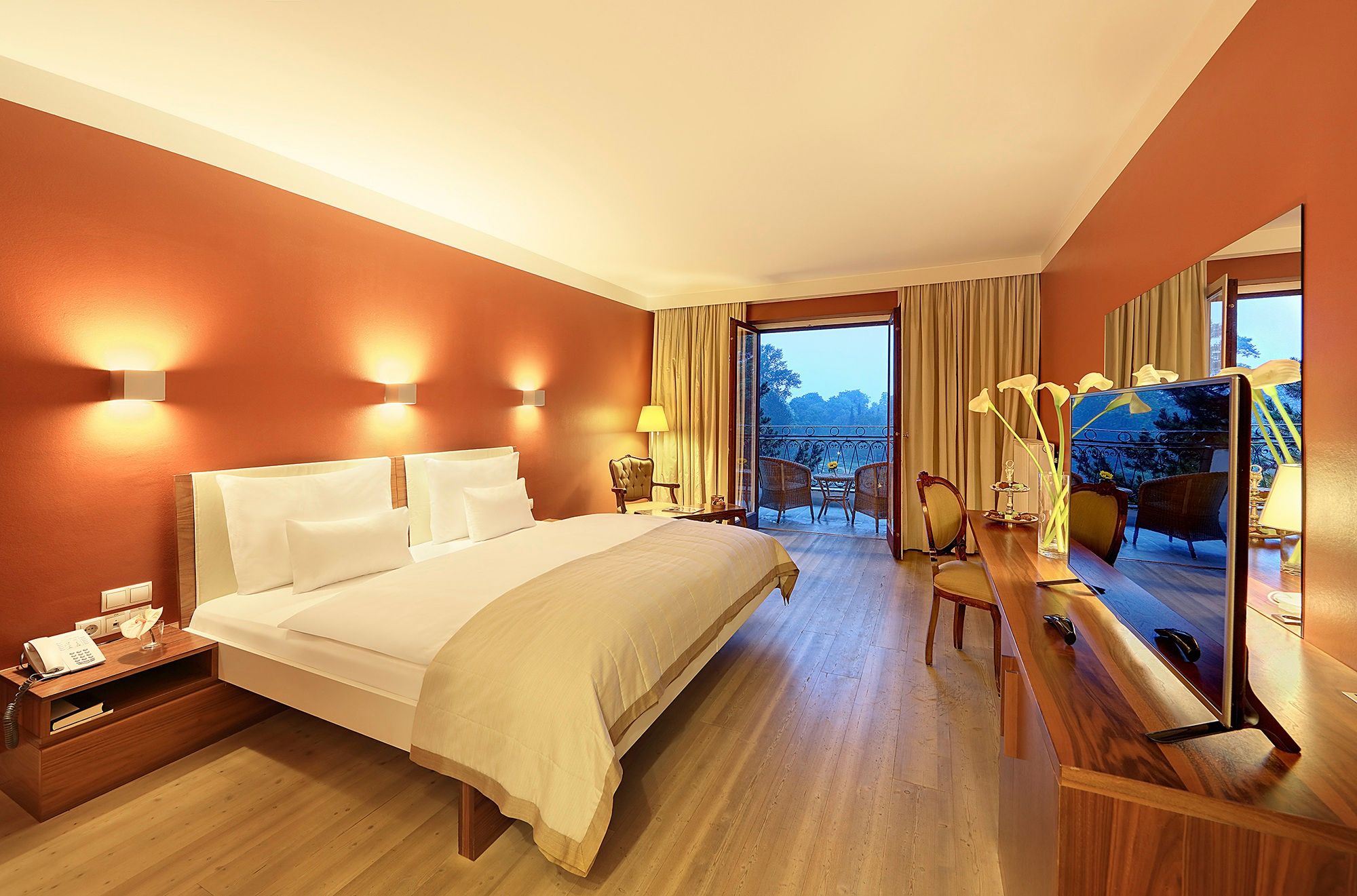 A modern hotel room with a double bed, warm wall colors, a balcony, and a view of the greenery.