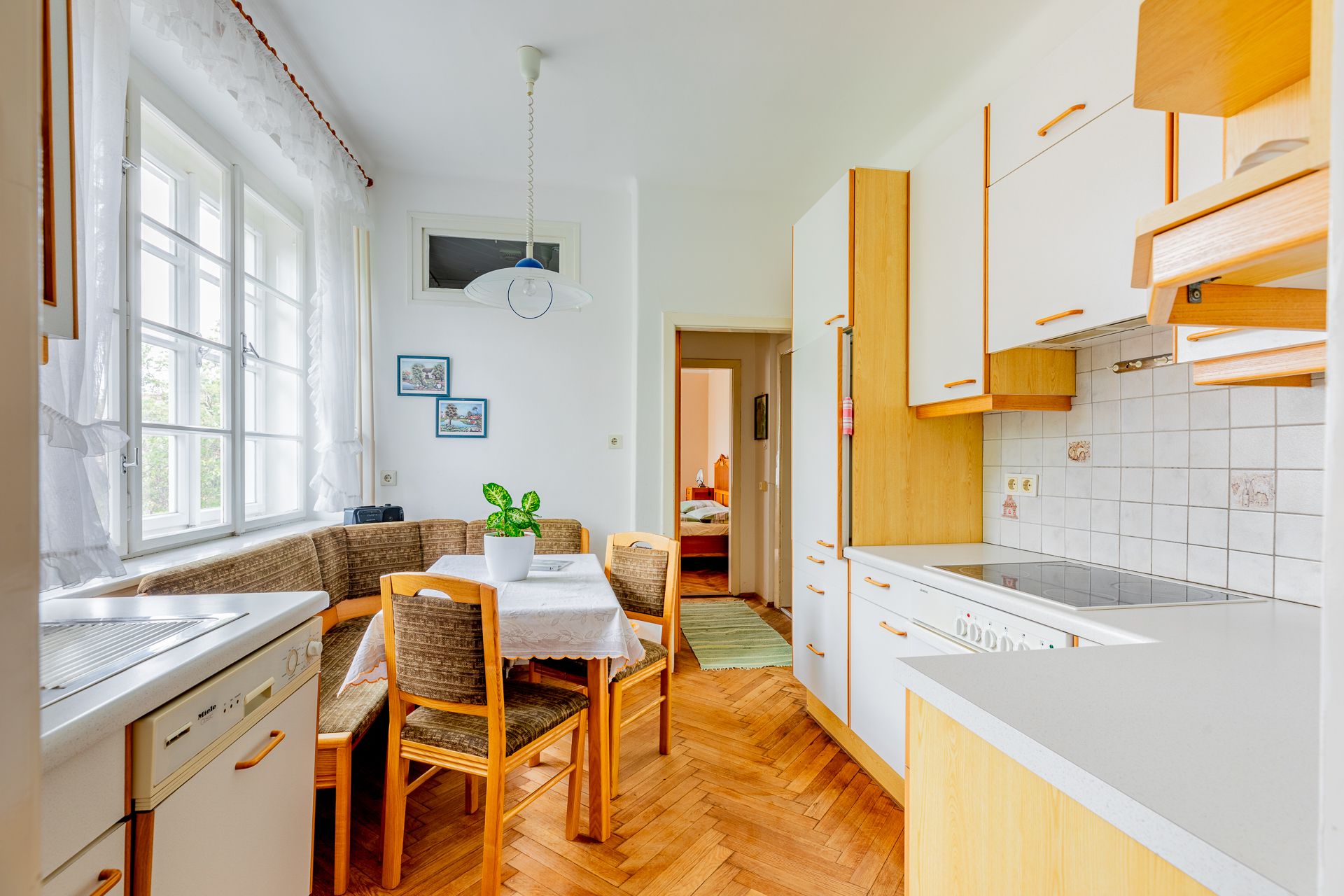 Dining area with wooden furniture, corner bench, table and chairs, bright kitchen with window, plants on the table.