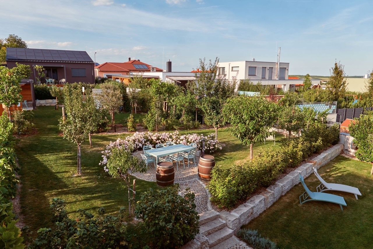A well-tended garden with trees, flowers, a table with chairs and two sun loungers. Modern houses can be seen in the background.