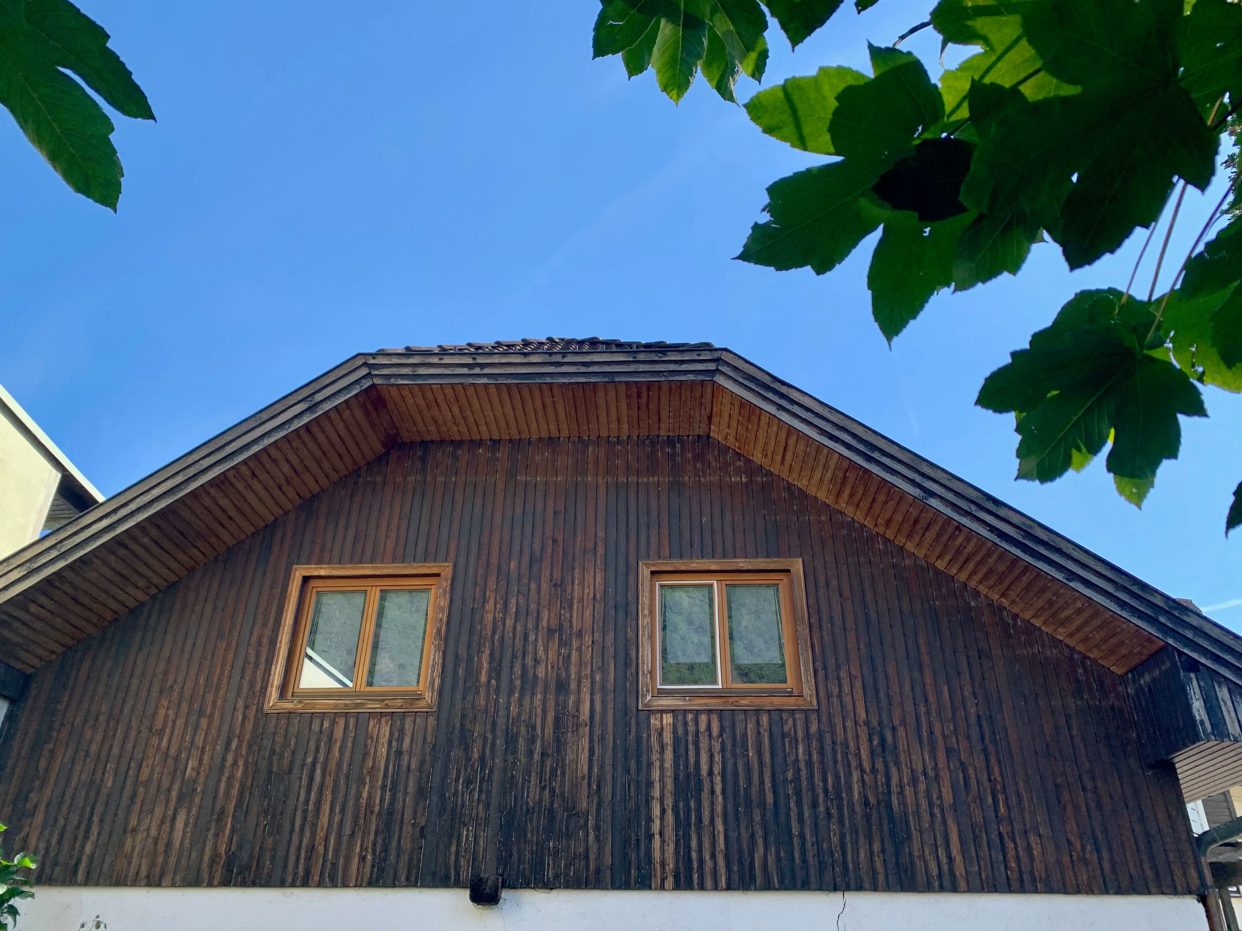 Attic of a house with wooden paneling and two windows, surrounded by green leaves and blue sky.