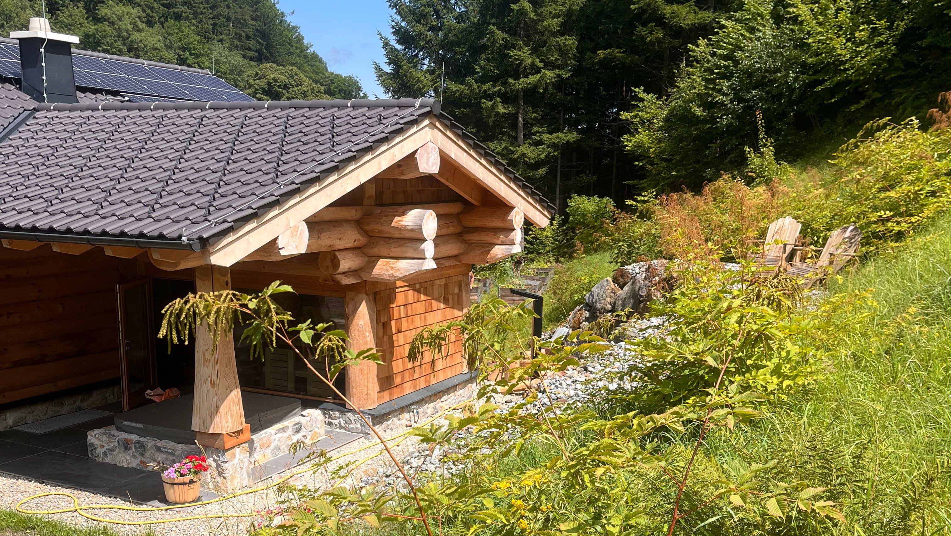Wooden chalet with solar panels on the roof, surrounded by forest and meadow.