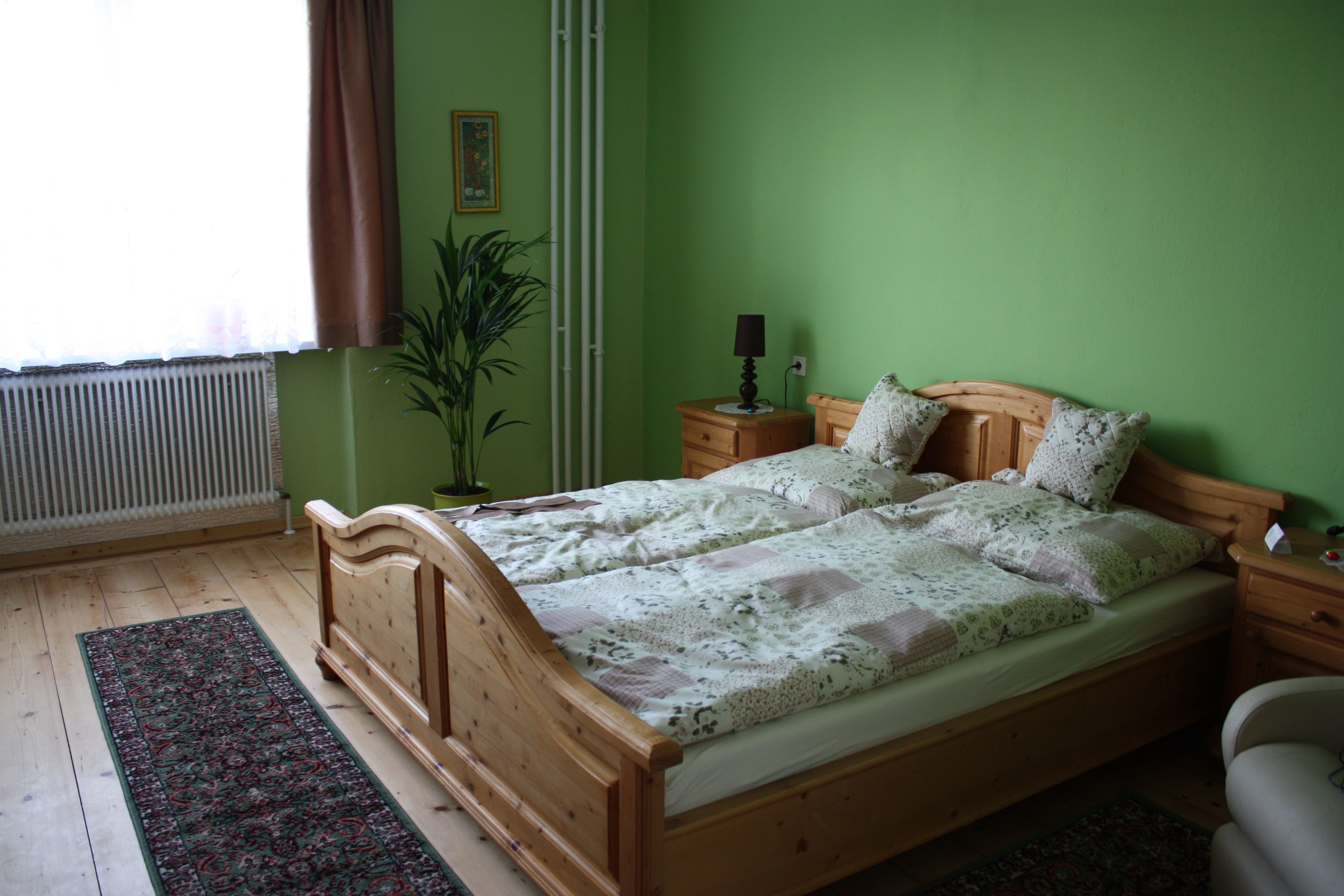 A cozy guest room with green-painted walls, a wooden bed, bedside tables, a plant and a carpet on a wooden floor.