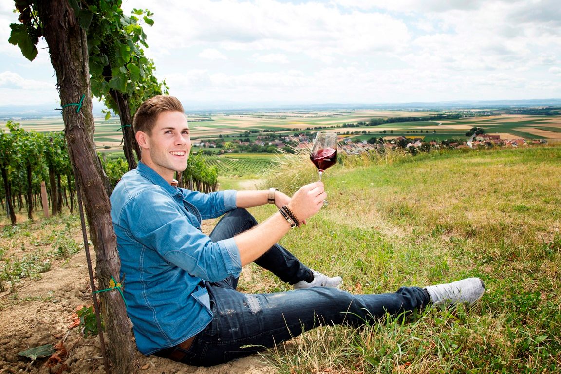 A man sits in a vineyard holding a glass of red wine. A vast landscape can be seen in the background.