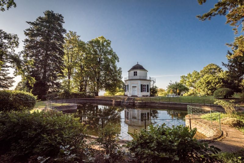 A round pavilion stands on the banks of a small pond, surrounded by trees and a well-tended garden.