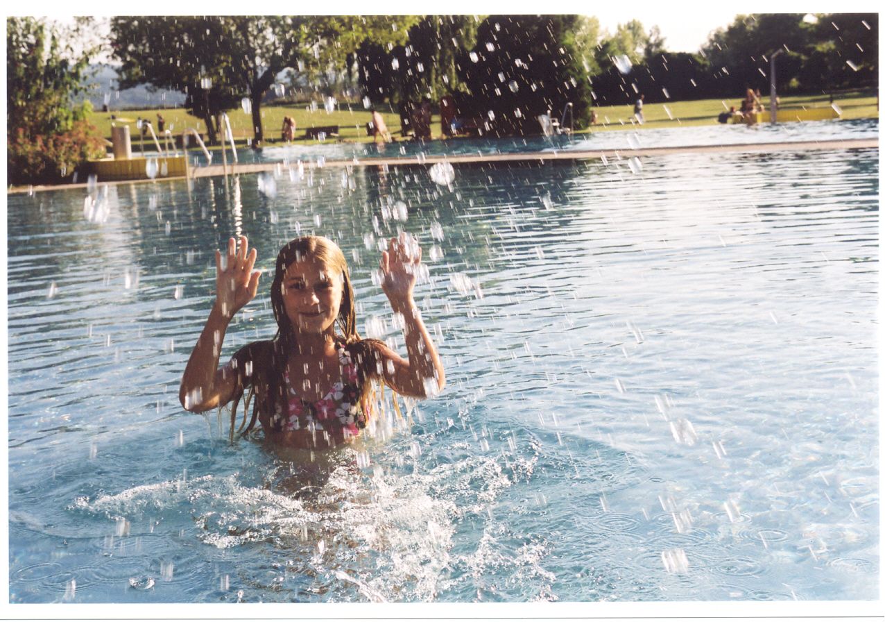 A girl in the water of an outdoor pool, surrounded by splashing water, with trees and meadow in the background.