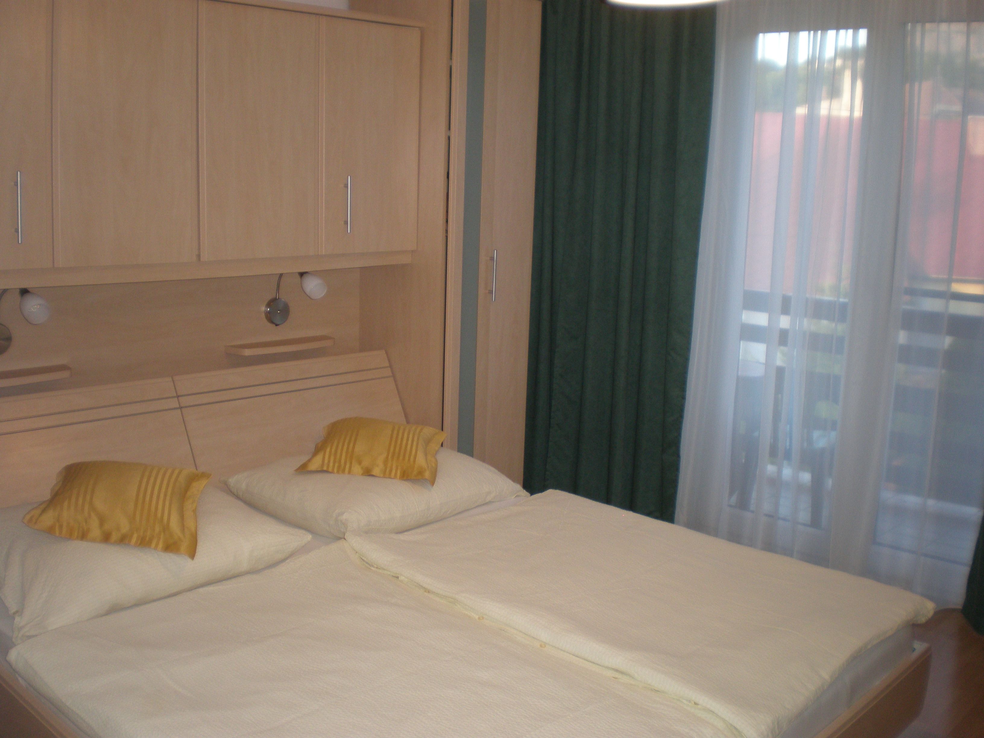 A bedroom with a double bed, yellow cushions, wooden wardrobes and a window with green curtains.