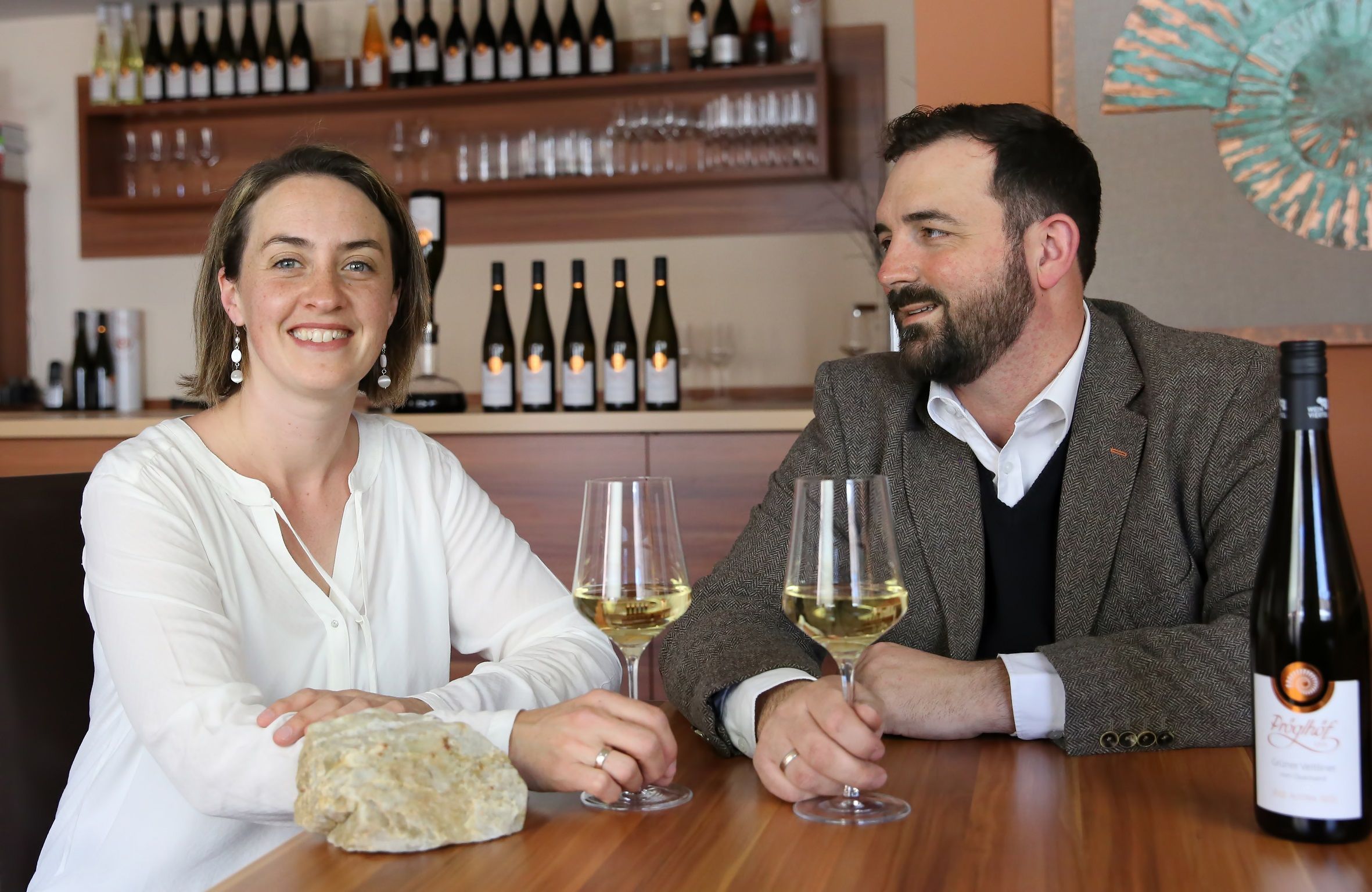 Two people are sitting at a table with wine glasses and bottles of wine in the background.