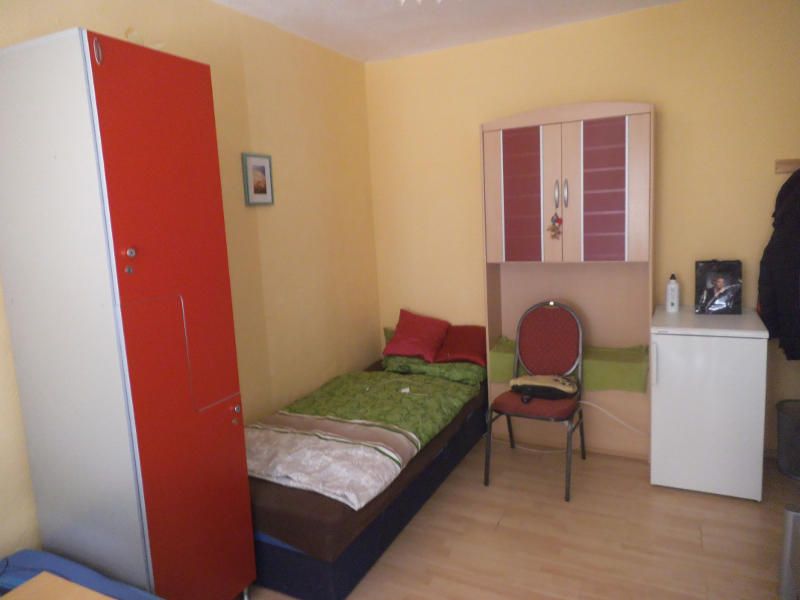 A small single room with a bed, red wardrobe, chair and small fridge.